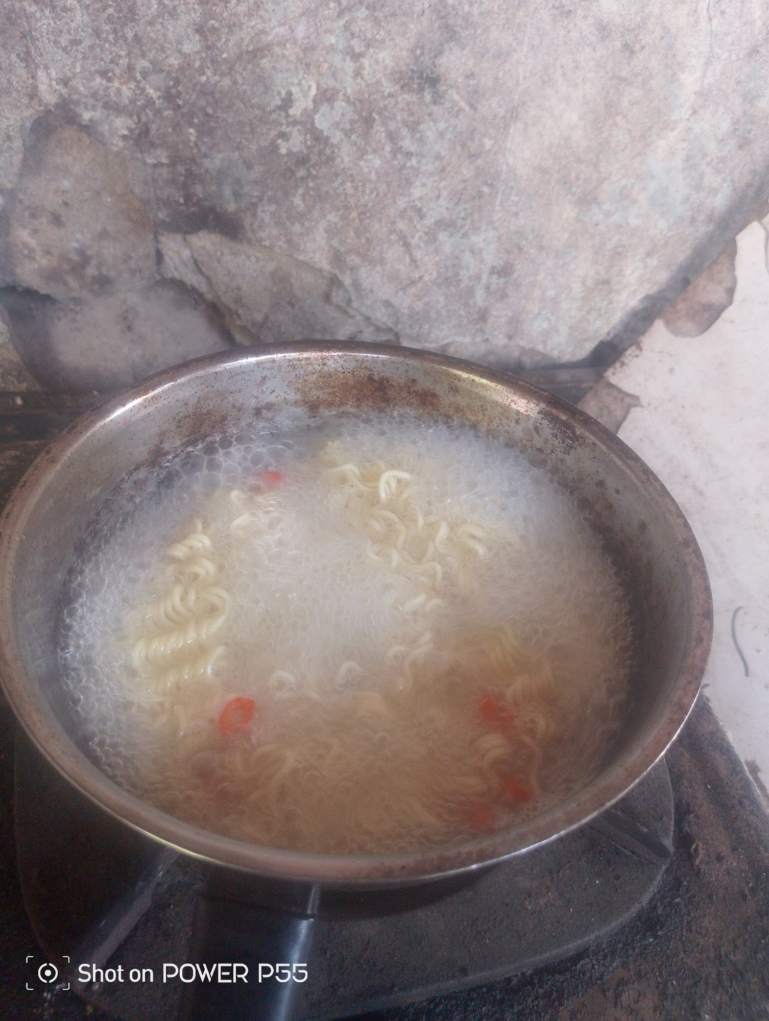 A close-up, eye-level shot shows instant noodles boiling in a metal pot on a stove. The pot, which appears old and slightly discolored, sits on a dark, textured stovetop. The noodles are a pale yellow and are submerged in a frothy, bubbling liquid. Small red flecks, possibly chili or seasoning, are visible within the noodles. The background is out of focus, but suggests a rustic setting with a large, rough, grey stone structure and a lighter, possibly concrete, surface. A watermark in the bottom left corner reads "Shot on POWER P55". The scene likely depicts a simple meal preparation.