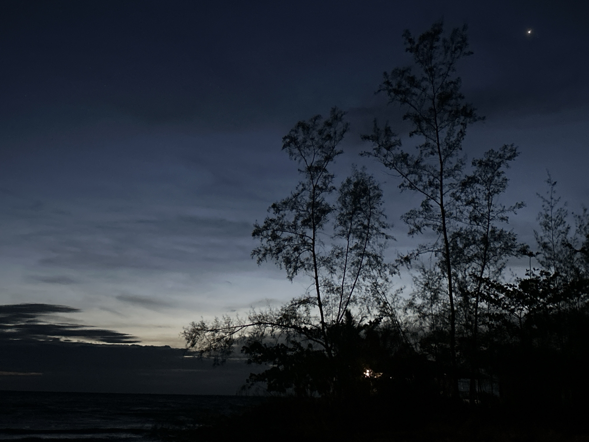 The image depicts a nighttime scene at a beach near Baan Sri Thanu, Thailand. The sky is a gradient of dark blue to light gray near the horizon, with clouds present. A crescent moon is visible in the upper right of the image. Silhouetted trees are present in the midground and foreground. The ocean is visible in the lower left, with small waves. No people are visible. A light source is emanating from the ground, located in the darkness.