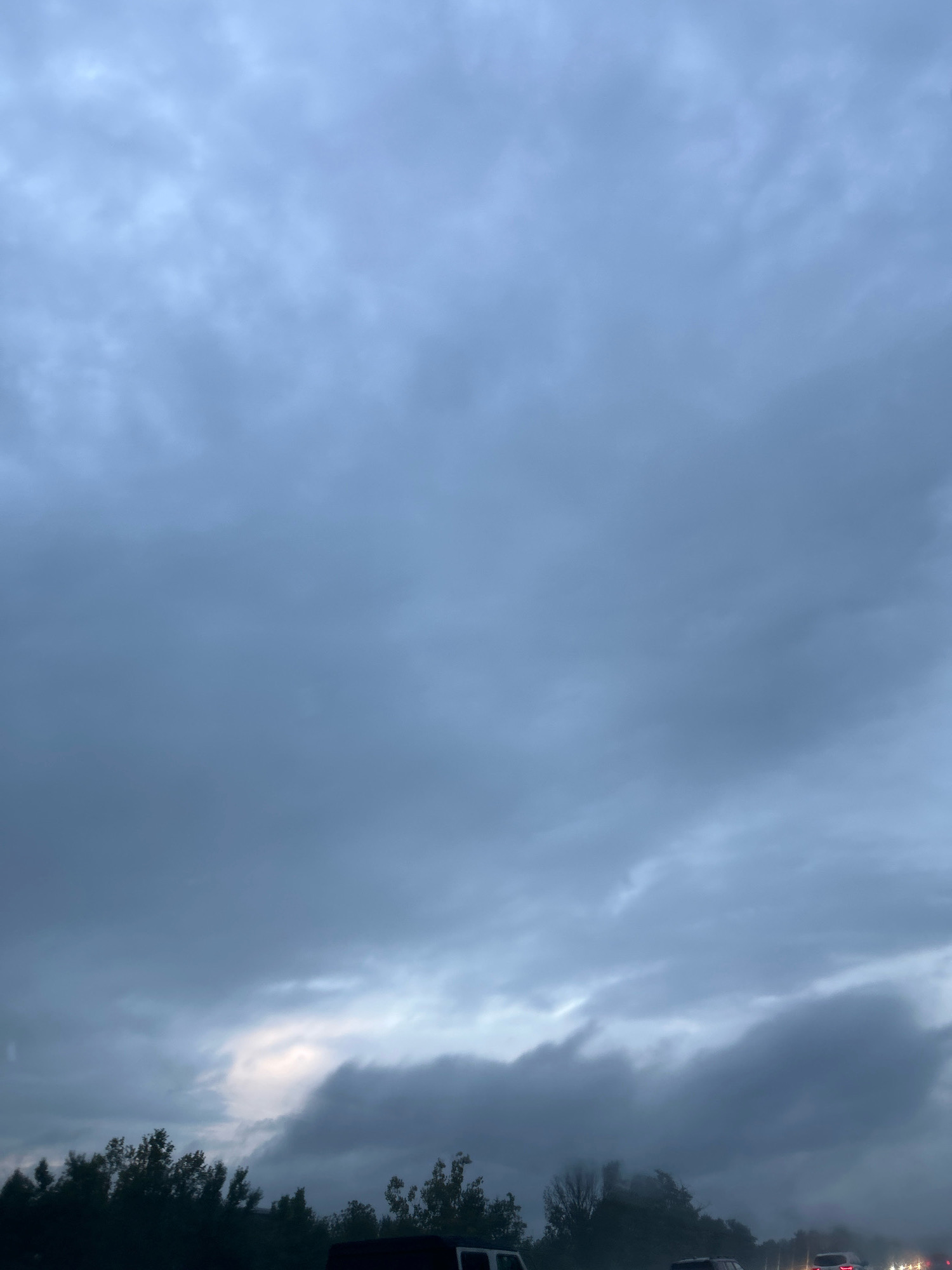 This is an eye-level, medium shot photograph taken from within a vehicle on a highway in Collier Township, United States of America. The sky is overcast with a heavy, dark grey cloud cover, and there is a faint break in the clouds with a patch of lighter sky towards the horizon. The bottom of the frame shows the dark silhouettes of trees and other vehicles on the road. The overall lighting is dim, suggesting it is either early morning or late evening, or that the weather is poor.
