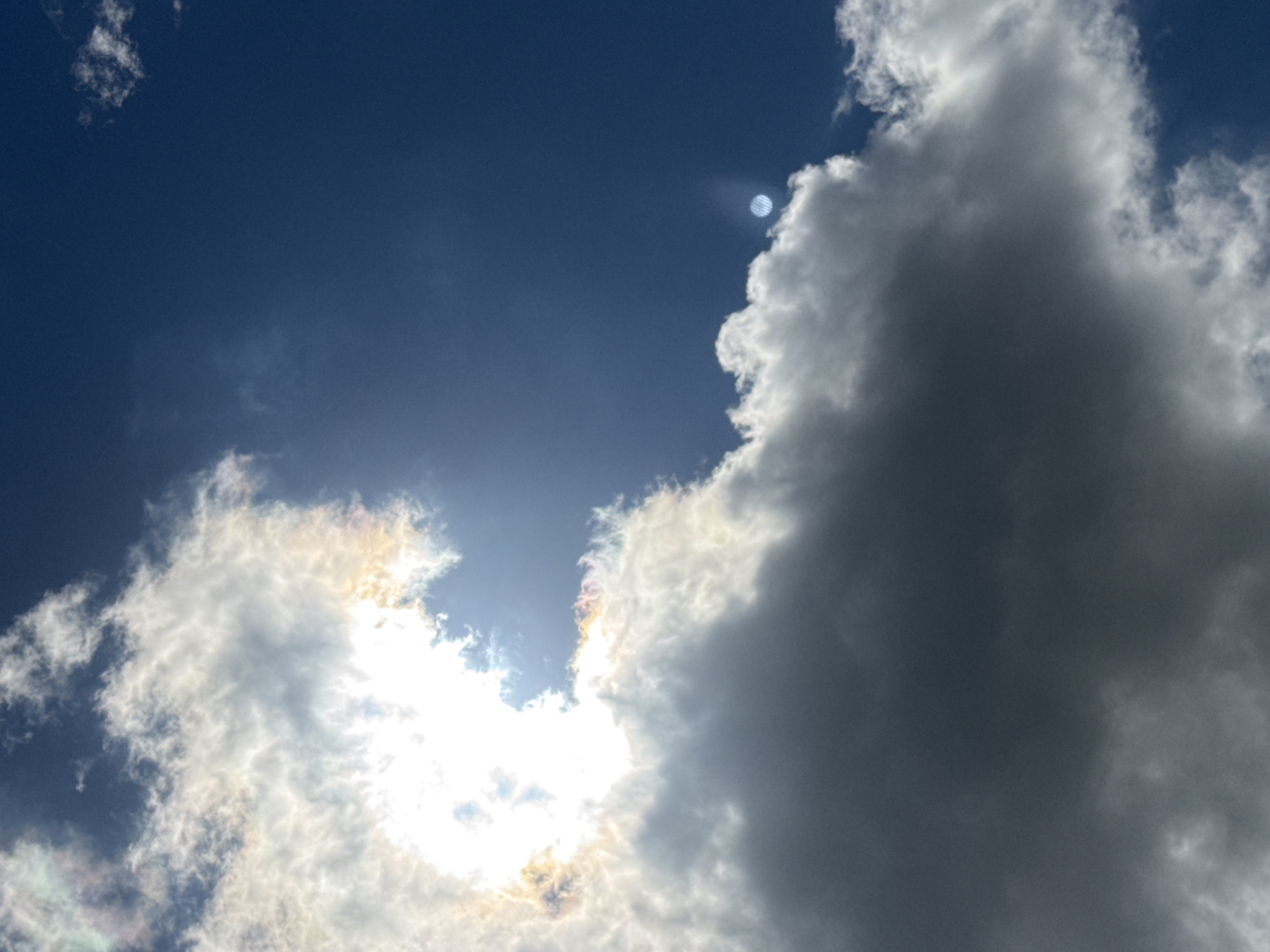 This image depicts a sky filled with a dramatic interplay of sunlight and clouds. The sun is partially obscured by thick, bright white clouds that are tinged with iridescent colors, possibly indicating a solar halo or iridescence within the clouds themselves. Below these, darker, heavier clouds loom, suggesting a change in weather or the presence of storm clouds. The sky is a deep, clear blue, contrasting with the varied cloud formations. There is a faint, circular lens flare or atmospheric phenomenon visible in the upper right quadrant of the sky. The scene is devoid of any urban or natural landscape elements, focusing solely on the sky and its atmospheric conditions. It is not possible to determine the location in the city of Asha, Russia from the provided image.