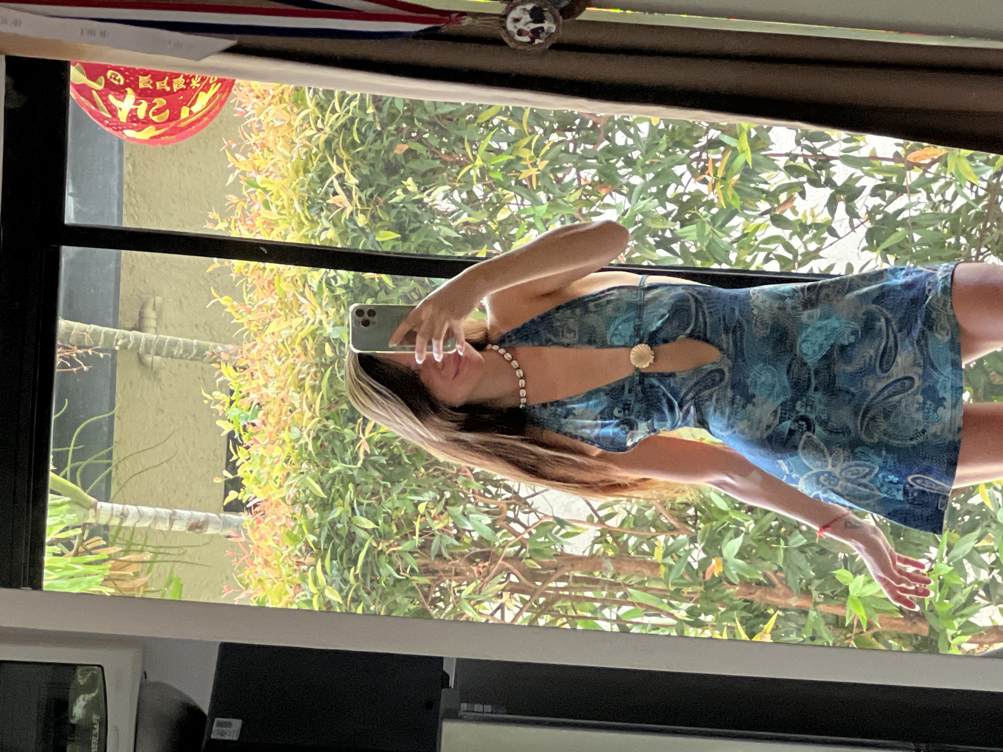 In an indoor setting, a young woman with long, brown hair is taking a selfie in a mirror. She is wearing a blue, paisley-patterned halter dress with a plunging neckline and a shell-shaped clasp. A pearl necklace is visible around her neck. Her left arm is raised, holding a smartphone. Her right arm is extended downwards, with her hand open. She is wearing a thin red bracelet on her left wrist. The background visible through a window consists of lush green foliage and a palm tree. A red, decorative lantern with Chinese characters is hanging in the upper right portion of the frame. A brown curtain is drawn partially on the right side of the image. There are also some items on a shelf to the left of the frame.