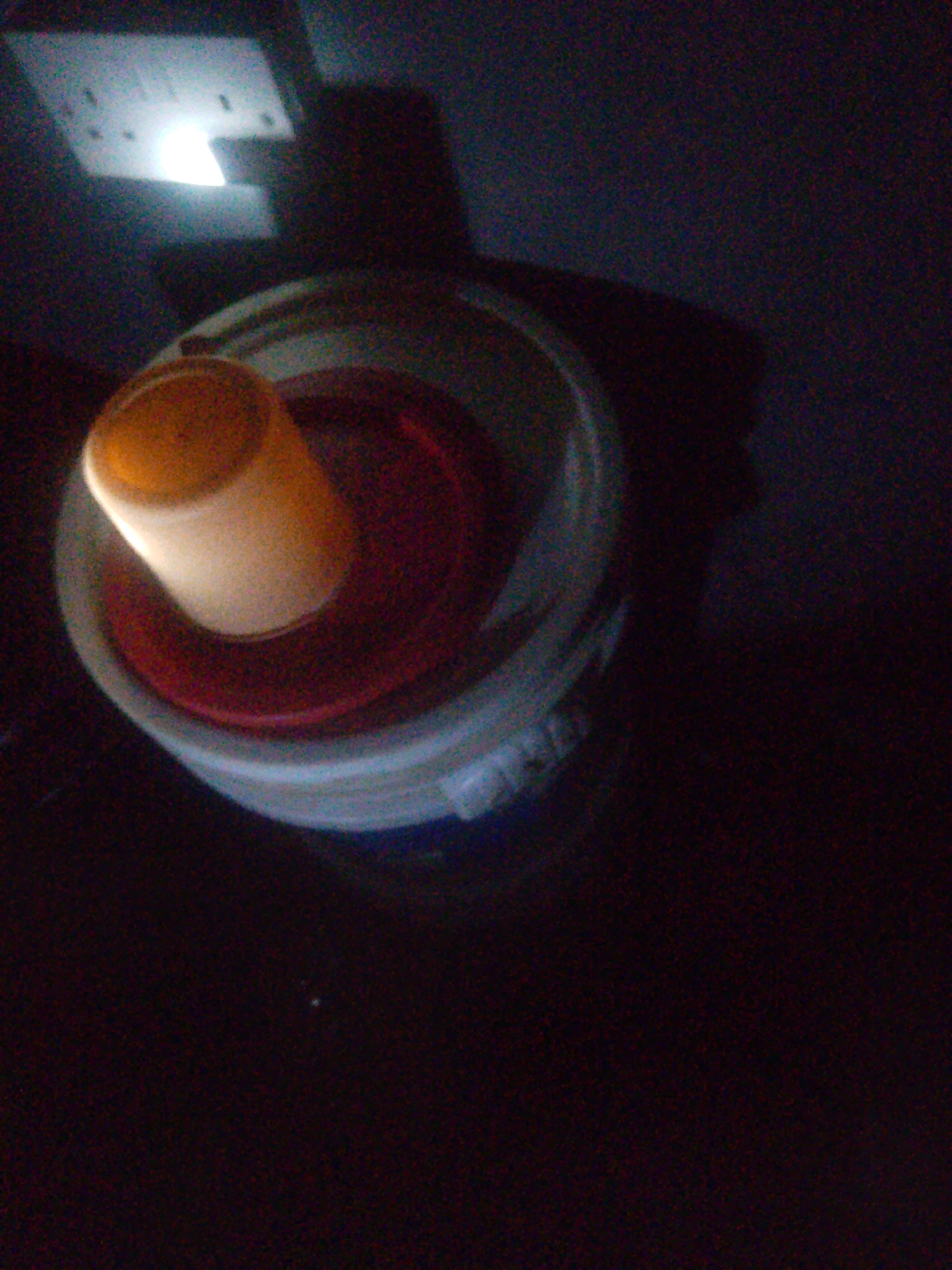The image depicts a close-up, overhead view of a cylindrical container, possibly a bucket, with a reddish-orange substance filling its lower portion. A lighter-colored, orange cylindrical object is partially submerged in this substance. The container itself has a greyish-white rim with some subtle blue-green reflections, suggesting it may be plastic. The background is dark and appears to be an indoor environment, with a faintly illuminated rectangular object in the upper left corner, possibly a light fixture or a screen. The overall lighting is dim, and the image has a grainy texture, indicative of low-light photography. There is no visible indication of the specific location within Jalingo, Nigeria, or any people, actions, or interactions.