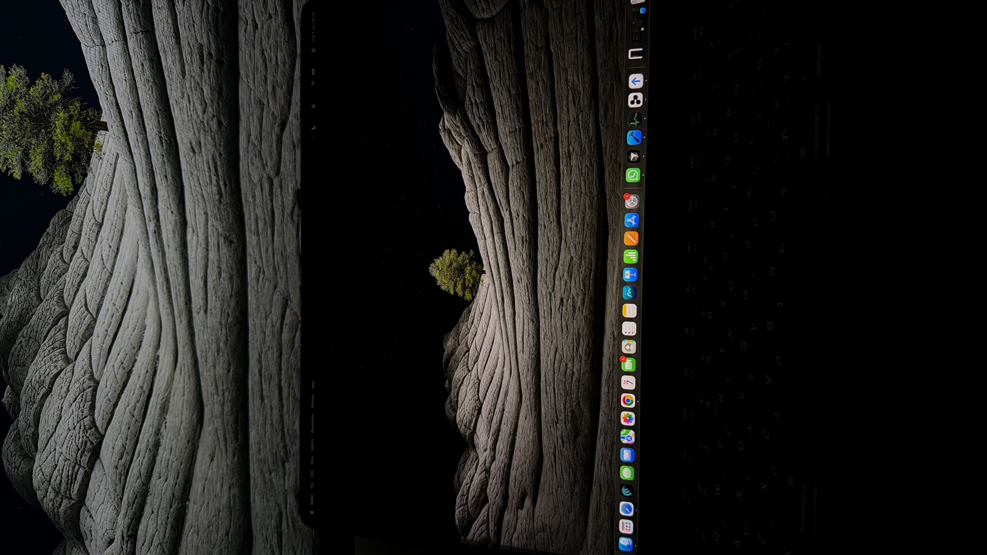 The image shows a split view of a landscape. The top portion depicts a close-up of a textured rock formation under a dark sky with a single tree visible in the upper right corner. The bottom portion shows a laptop screen displaying the same landscape as a wallpaper. The laptop screen also shows a dock with application icons and a menu bar at the top. The date and time "Tue 7 Jul 19:19" are visible in the top right corner of the screen. The image is not specific to Kafr Yasif, Israel.