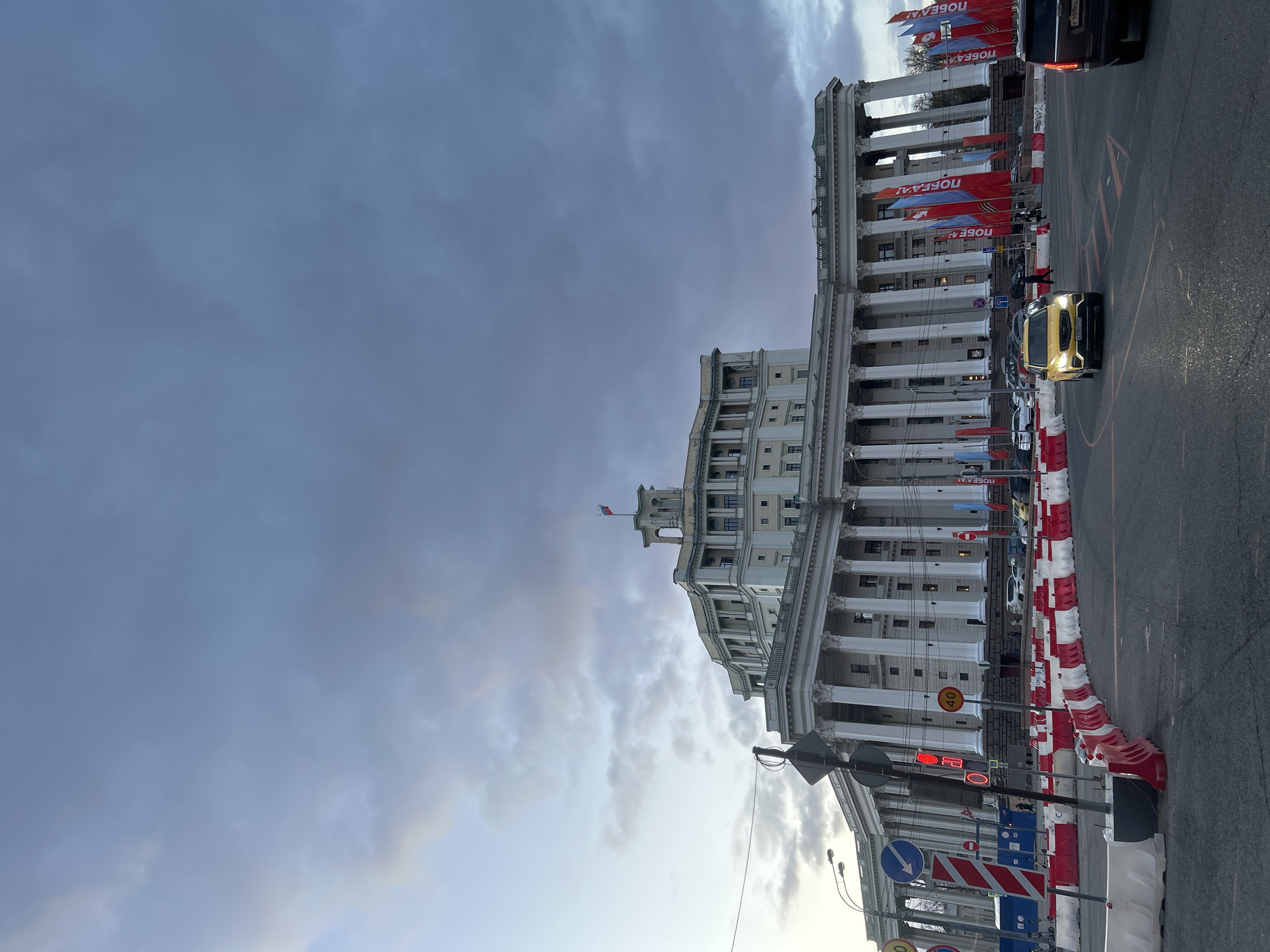 The image displays a grand, classical-style building lining a city street in Moscow, Russia, under an overcast sky. The large, multi-story structure, possibly an administrative building, features numerous tall columns along its facade and a small domed or cupola-like structure at its top, where a flag, likely the Russian flag, is flying.
In the foreground and mid-ground, the street is partially cordoned off by red and white striped traffic barriers. Several vehicles are visible, including a prominent yellow SUV with its headlights illuminated, driving forward. Other cars are in traffic or parked nearby. Various street signs are present, such as a "40" speed limit sign, blue directional signs, and a red traffic light.
A striking detail is the series of red banners adorned with white text, hanging vertically between the columns of the building. The visible text on these banners reads "ПОБЕДА!" (Victory!), strongly suggesting a celebration or commemoration, likely related to Victory Day. The sky is predominantly cloudy, with some lighter patches indicating either an overcast daytime or the approach of dawn or dusk. The scene depicts a busy urban setting prepared for a patriotic event.