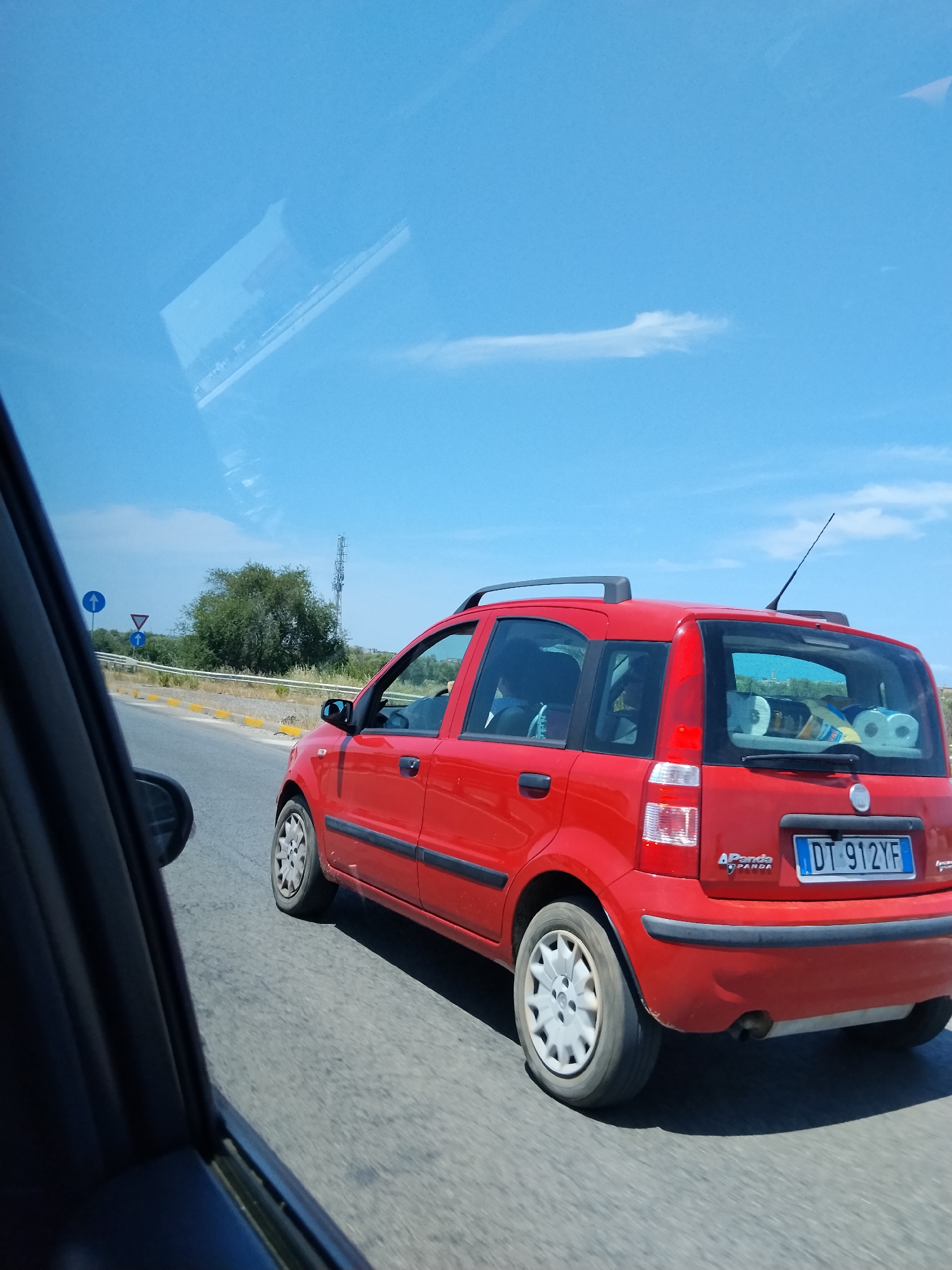 The image depicts a roadside scene in Bisceglie, Italy, observed from the front passenger perspective of a moving vehicle. A red, 2nd generation Fiat Panda, bearing the Italian license plate "DT 912YF", travels alongside and slightly ahead to the right of the observer. The red vehicle is fitted with a black roof rack. The rear cargo area is visibly occupied by several rolls of toilet paper and other miscellaneous items. A driver is visible through the front driver's side window. The asphalt road surface features a white guardrail and yellow-painted curbs on the left. Three road signs are present on the left: two blue circular signs indicating "straight ahead" (compulsory direction) and one red triangular "yield" sign. Behind the guardrail, a dense line of green trees and bushes flanks the road. In the distance, a tall cellular communication tower is visible against a clear, bright blue sky with sparse white clouds. Both vehicles appear to be in motion under daylight conditions.