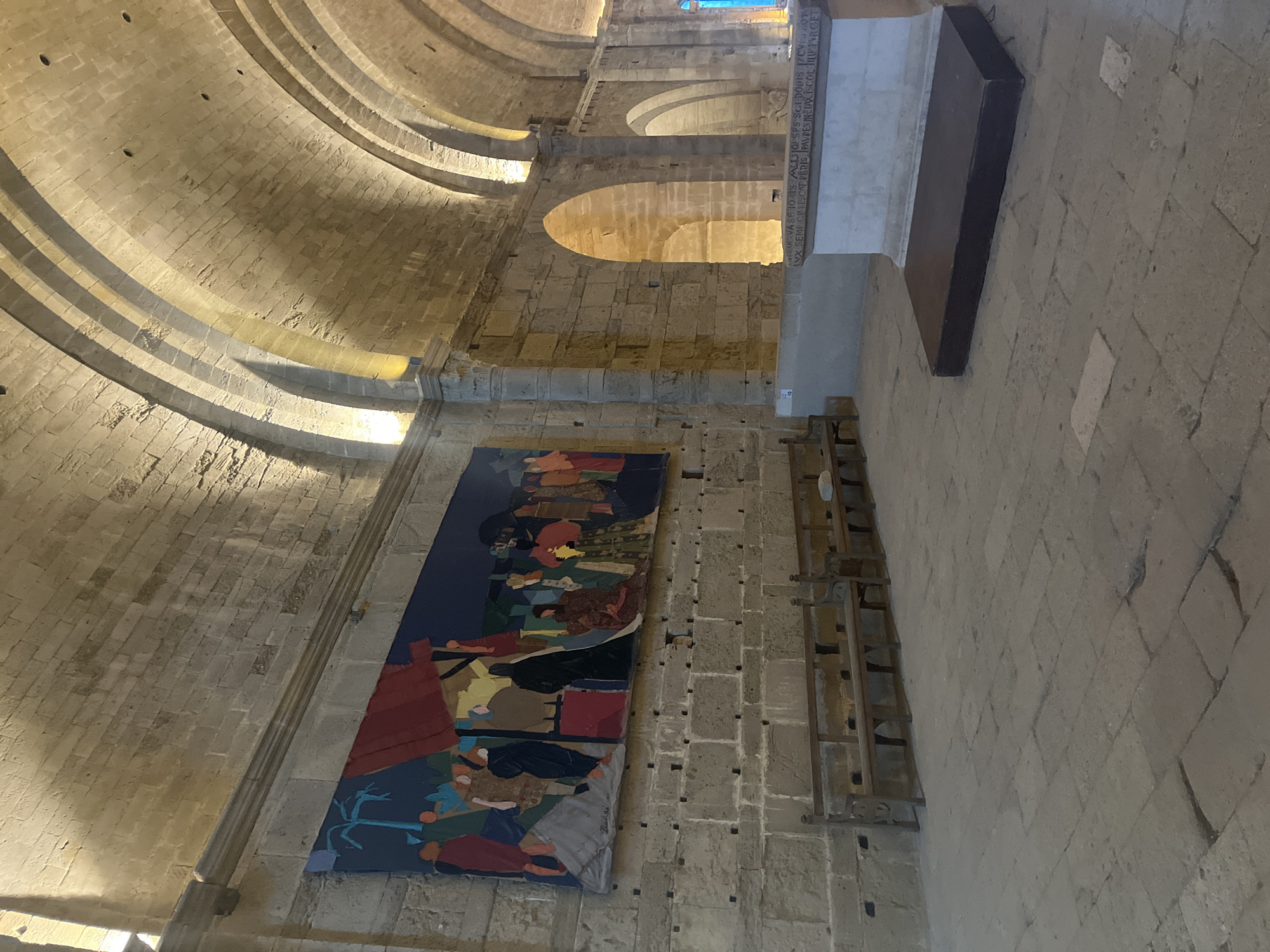 The image shows the interior of the Cathédrale Saint-Pierre de Villeneuve-lès-Maguelone in Villeneuve-lès-Maguelone, France. The stone structure features high, arched ceilings and massive stone walls. A large, colorful tapestry depicting a crowded scene with figures in robes hangs prominently on the left wall. In the foreground, there are several rustic wooden benches lined up. To the right, a large stone altar or monument is visible, with a dark rectangular platform in front of it. The floor is paved with large, uneven stone tiles. The lighting is warm and appears to be artificial, highlighting the texture of the stone and the artwork.
