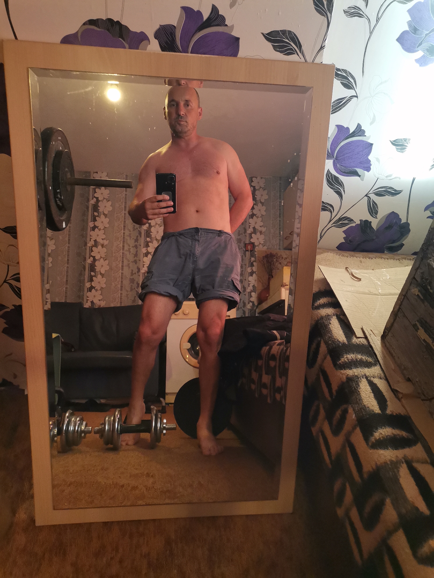 A shirtless man with a shaved head is depicted taking a mirror selfie in an indoor room. He wears grey cargo shorts and holds a black smartphone to capture his reflection. The man stands with his feet apart and hands clasped behind his back, showcasing his torso and legs.
The large mirror, framed in light-colored wood, reflects the man and the room's interior. In the reflection, a barbell with black weight plates is visible to the man's left. Behind his lower body, a dark-colored sofa and a white washing machine with a distinctive circular yellowish-orange door are reflected. On the light brown carpeted floor in front of the mirror, two dumbbells with metallic weights are placed.
The room's walls are adorned with patterned wallpaper. The section above the mirror features large purple and black floral designs on a light background. The wallpaper visible in the reflection behind the man has a different pattern of smaller white flowers on a darker background. A prominent bright light source is reflected in the upper left portion of the mirror. To the right of the mirror's frame, part of a brown and black patterned rug or blanket, and a piece of cardboard or wood, are visible. The overall scene suggests a personal, multi-functional space, likely within a home, possibly serving as a home gym or utility area. The man's expression is neutral. No text is visible in the image.