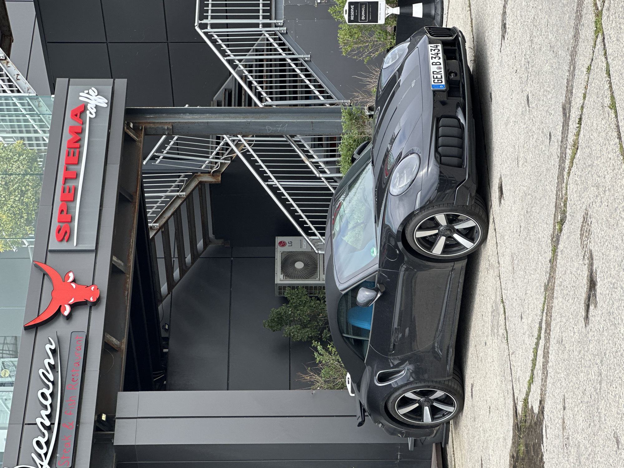 An image, rotated 90 degrees clockwise, captures a black sports car parked alongside a modern building that houses a restaurant and cafe. The scene suggests an urban setting during what appears to be daytime under an overcast sky.
The prominent subject is a dark grey or black Porsche 911 convertible, possibly a 992 generation, parked on a light grey paved surface with visible cracks and some grass growing through them. The car features multi-spoke, two-tone wheels and a light blue interior, visible through the windshield. Its front license plate, "GER B 3434", indicates a German registration.
The building to the left of the car (when upright) is dark grey with a minimalist, panelled facade. A large sign for "SPETEMA caffe" is mounted vertically, with "SPETEMA" in red capital letters and "caffe" in white cursive. Below this, a red stylized bull or ox head logo is visible, followed by white cursive text that reads "anarm" (partially visible) and "Steak & Fish Restaurant" in smaller red text on a dark background.
Behind the car, on the building's exterior, there are metal stairs and walkways, suggesting multiple levels or an industrial aesthetic. An LG air conditioning unit is mounted on the wall near the car's windshield. Further to the right, a small white sign with black text reads "RESERVE" at the top and "DUNHILL" below it, indicating a designated parking spot or area. The overall impression is a contemporary business establishment with a high-end car parked outside.