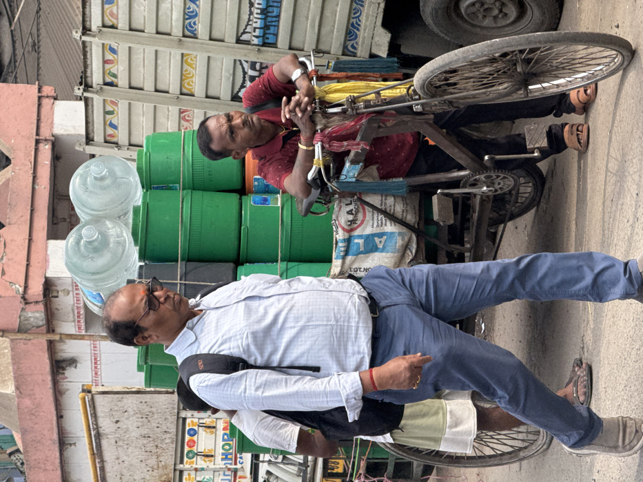 Two men are next to a cart bicycle carrying water bottles and green containers. One man in a red shirt is leaning on the bicycle while the other is standing next to the cart with a blue shirt and pants. The cart is parked next to a truck with a white panel. Buildings are in the background.