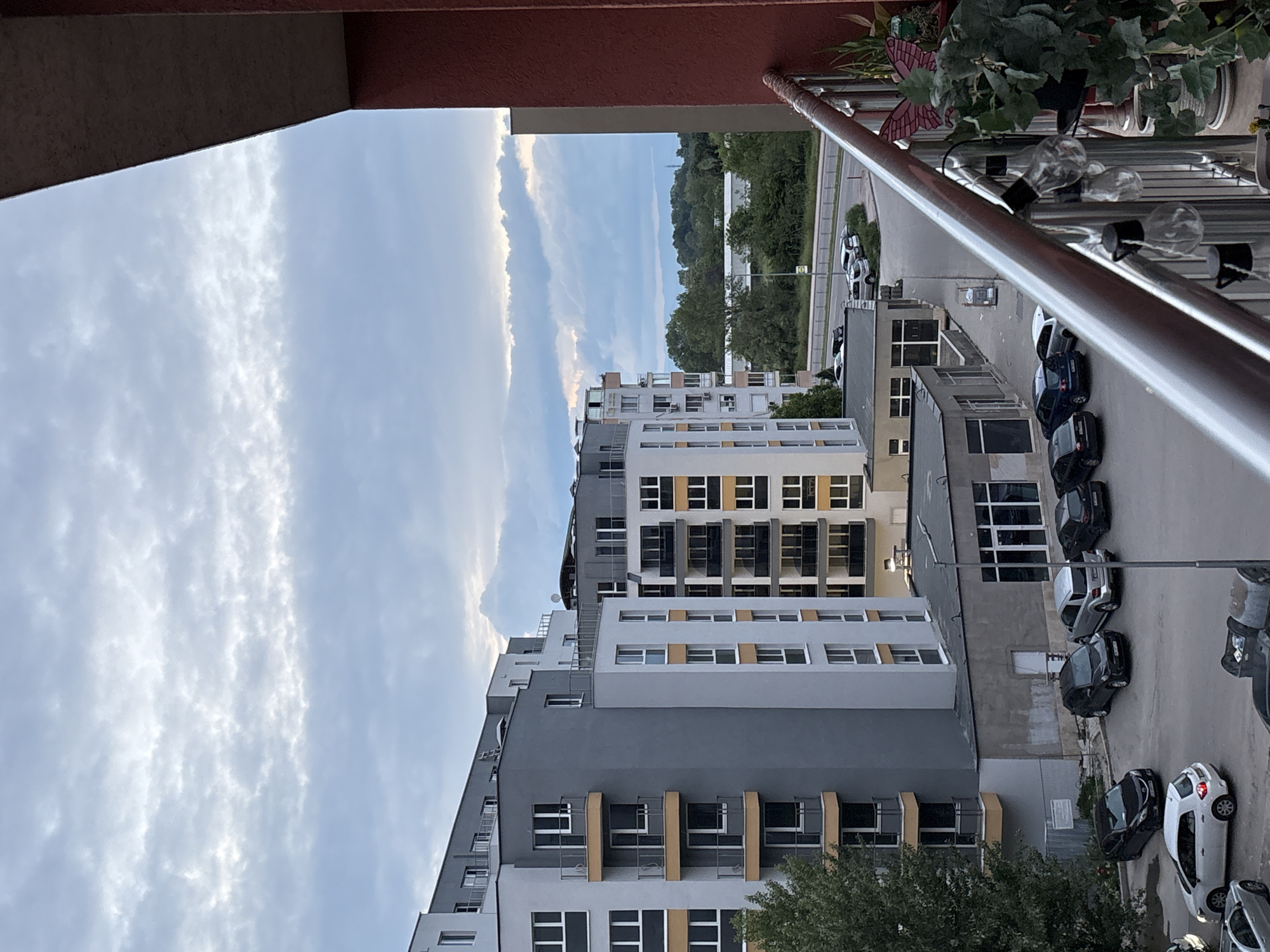 The image depicts an urban scene in Plovdiv, Bulgaria, viewed from a high vantage point, likely a balcony.
The focal point is a tall apartment building complex with a mix of gray and white facades, adorned with windows and balconies. A variety of parked cars line the street alongside the building, with a row of trees located nearby.
In the distance, one can see more trees and a glimpse of the city skyline. The sky overhead is partly cloudy.
In the foreground are elements of the balcony from which the photograph was taken, including a reddish-brown overhang, a metal railing, and a decorative light fixture with light bulbs and foliage.
