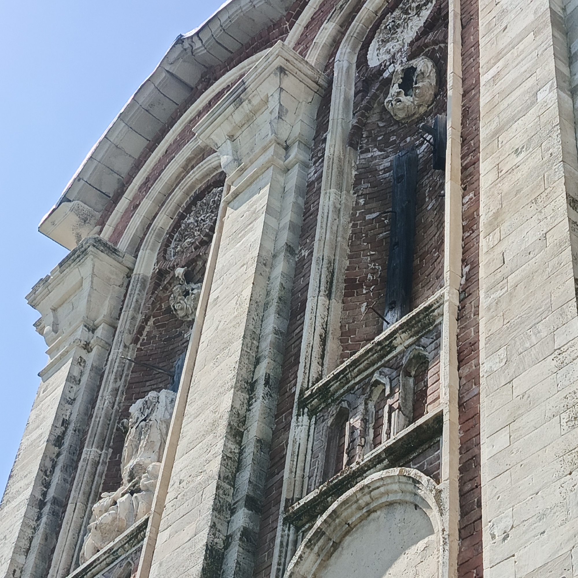 This is a low-angle, full shot of the facade of a historic building, likely a church or cathedral, under a clear blue sky. The architecture features alternating bands of light-colored stone and dark red brick. The stone elements are intricately carved with decorative molding and architectural details, including arched window frames and protruding decorative elements. There are at least two distinct arched niches visible. One niche, towards the top left, contains a damaged decorative carving resembling a human face or mask, with a dark, hollowed-out opening. Below this, another niche on the left contains a weathered, light-colored sculpture of human figures in a group. The building shows signs of age and weathering, with visible cracks in the stone and some moss or green growth on the decorative elements. The overall impression is of a grand, old structure. The image does not contain any people. The location of this building is the church of St. Nicholas in the village of Tugustemir, Bashkortostan, Russia.