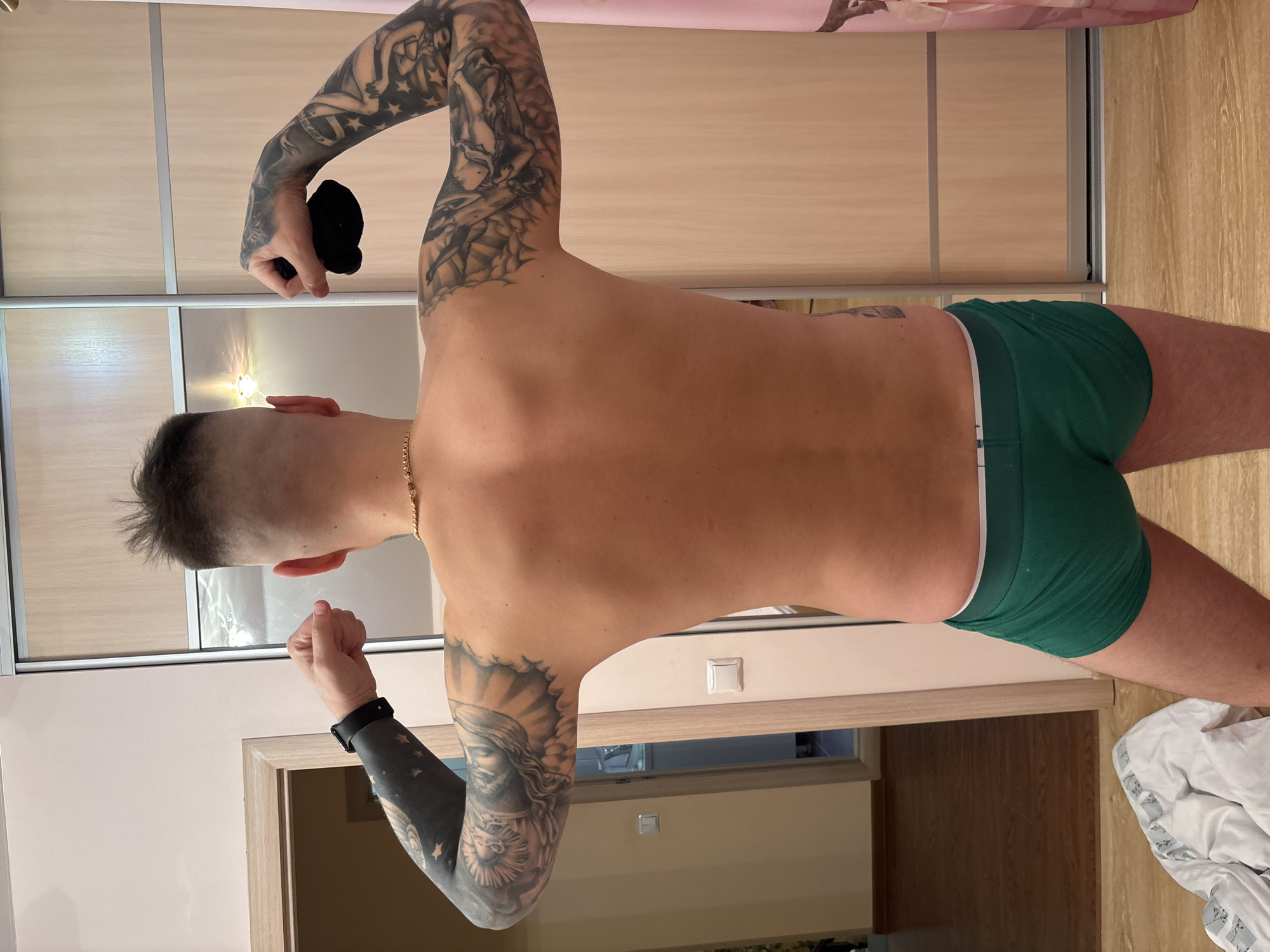 A male individual is depicted from the back, standing indoors and flexing both biceps. He has short, dark, faded hair and wears a thin gold-colored chain around his neck. His upper torso is bare, revealing a lean build. Both arms are heavily tattooed; the left arm features a mostly blacked-out forearm with white stars and detailed imagery on the upper arm, along with a black smartwatch on the wrist. The right arm is also fully tattooed with intricate designs, and the hand is clenched, holding a small, crumpled black fabric object. He is wearing green boxer briefs with a white waistband. The environment appears to be a residential room with light wooden flooring. To the left, a light-colored wall features a wooden door frame, beyond which another room with darker flooring is visible. Two white electrical switches are mounted on this wall. To the right, a large, light-colored wooden sliding wardrobe, which is partially reflective, occupies the background. A pink curtain or fabric is partially visible on the far right. A crumpled white fabric item rests on the floor in the lower-left foreground. The specific location within the city of Русский Вожой, Russia, cannot be determined from the visual information provided.