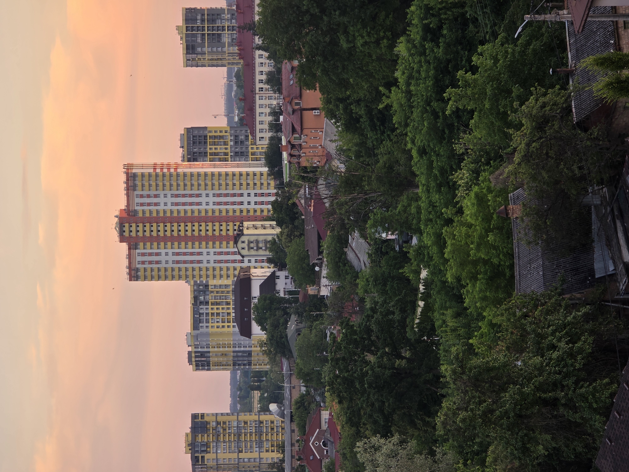 This is a vertical, slightly tilted, full shot of a cityscape at dusk in Kyiv, Ukraine. The sky is a soft gradient of pink and orange, indicating the sun has recently set or is about to rise. Tall, modern apartment buildings with colorful facades of yellow, red, and white dominate the left side of the frame. These buildings are interspersed with clusters of green trees, some of which are quite tall and dense, creating a contrast between the urban development and nature.
Below the apartment buildings, there are older, lower-rise buildings with red-tiled roofs and some brick structures, suggesting a mix of architectural styles and ages within the city. Power lines and streetlights are visible, hinting at the functioning infrastructure of the area. The overall atmosphere is peaceful, with the soft sky colors and the quiet presence of the buildings and trees. There is no visible text in the image.