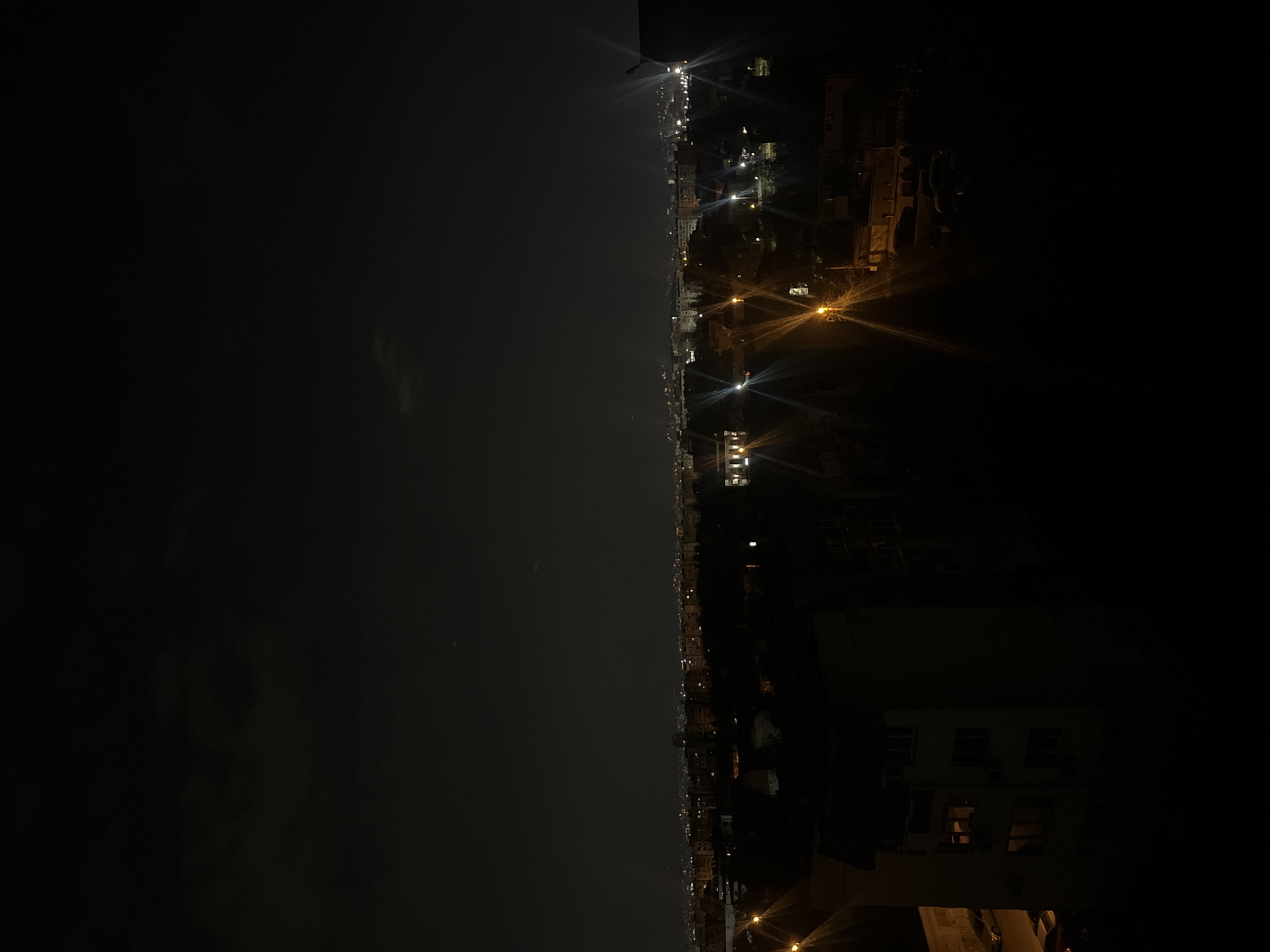 The image depicts a cityscape at night. The left side of the frame is predominantly dark, suggesting a body of water or open space. The right side shows a line of illuminated buildings and streetlights, indicating an urban area. The lights suggest Sofia, Bulgaria, as the city has a distinct nighttime skyline. The buildings range in height, and there are noticeable variations in light intensity and color, reflecting different types of illumination in the city. No people are identifiable in the scene. The image is taken from a high vantage point, possibly a tall building. The overall atmosphere is dark and still, punctuated by the glow of the city lights.