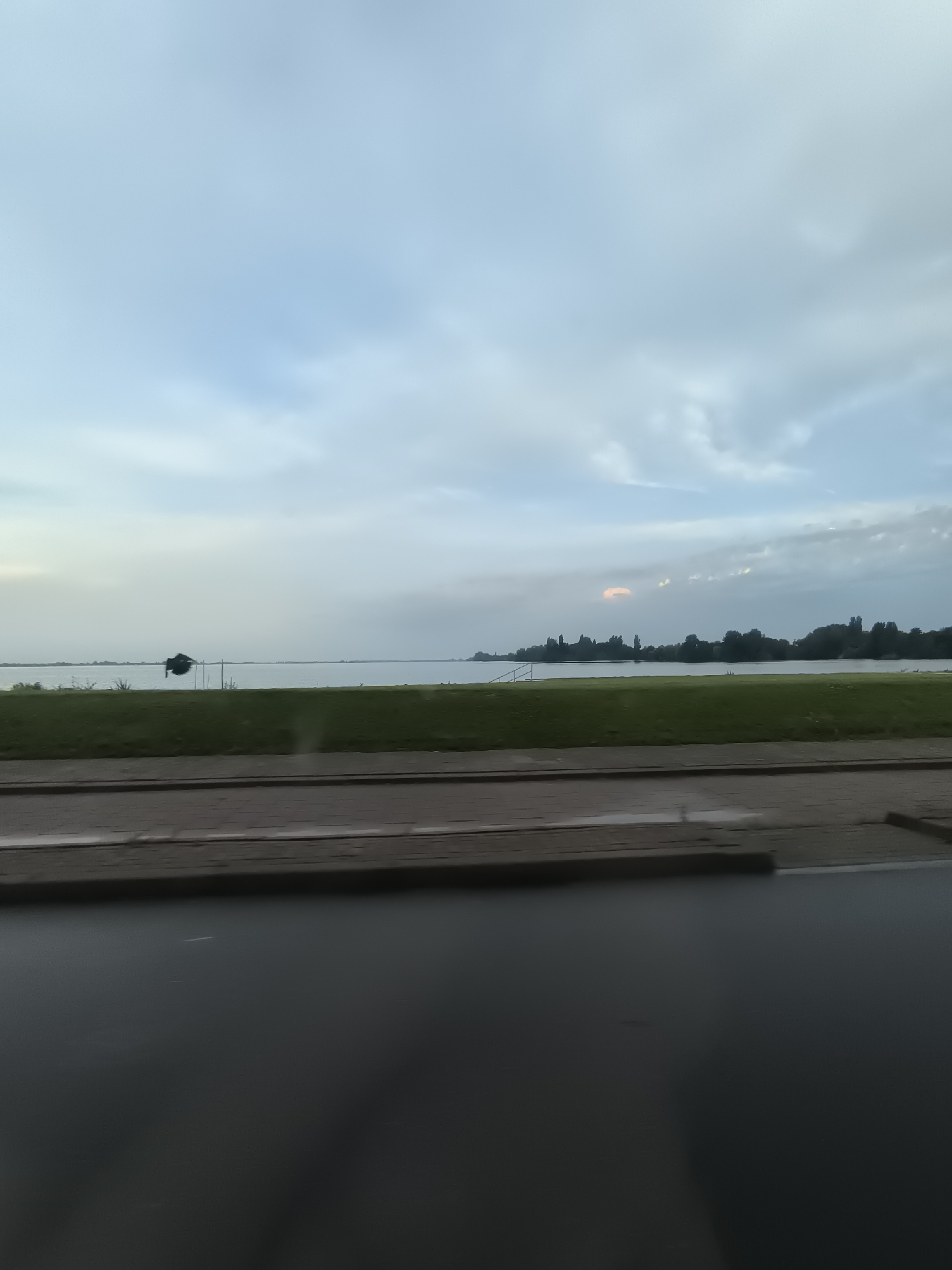 This image depicts a serene waterfront scene, likely taken from a moving vehicle, showcasing the tranquil waters of the Westeinderplassen in Aalsmeer, Netherlands. The foreground is dominated by a dark, reflective surface, possibly a road or pavement, followed by a strip of manicured green grass. Beyond the grass, the expansive Westeinderplassen stretches out, its surface calm and reflecting the overcast sky. A silhouetted object, possibly a small boat or structure, is visible on the water in the mid-ground, anchored by thin poles. The far bank of the lake is lined with a dense row of dark green trees, creating a natural silhouette against the horizon. The sky is a soft blue with diffused clouds, and a faint, warm-toned cloud formation is present in the distance, suggesting the possibility of a sunset or sunrise. The overall impression is one of quietude and natural beauty.