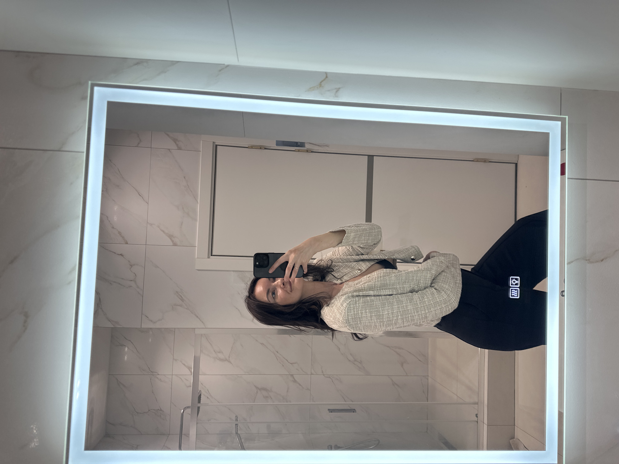 A person is seen in a mirror selfie. The person is wearing a light-colored tweed jacket and dark pants. The mirror has an illuminated frame. The background shows a bathroom with white marble-patterned tiles and a glass shower screen. The location is assumed to be Прикамский, Russia.