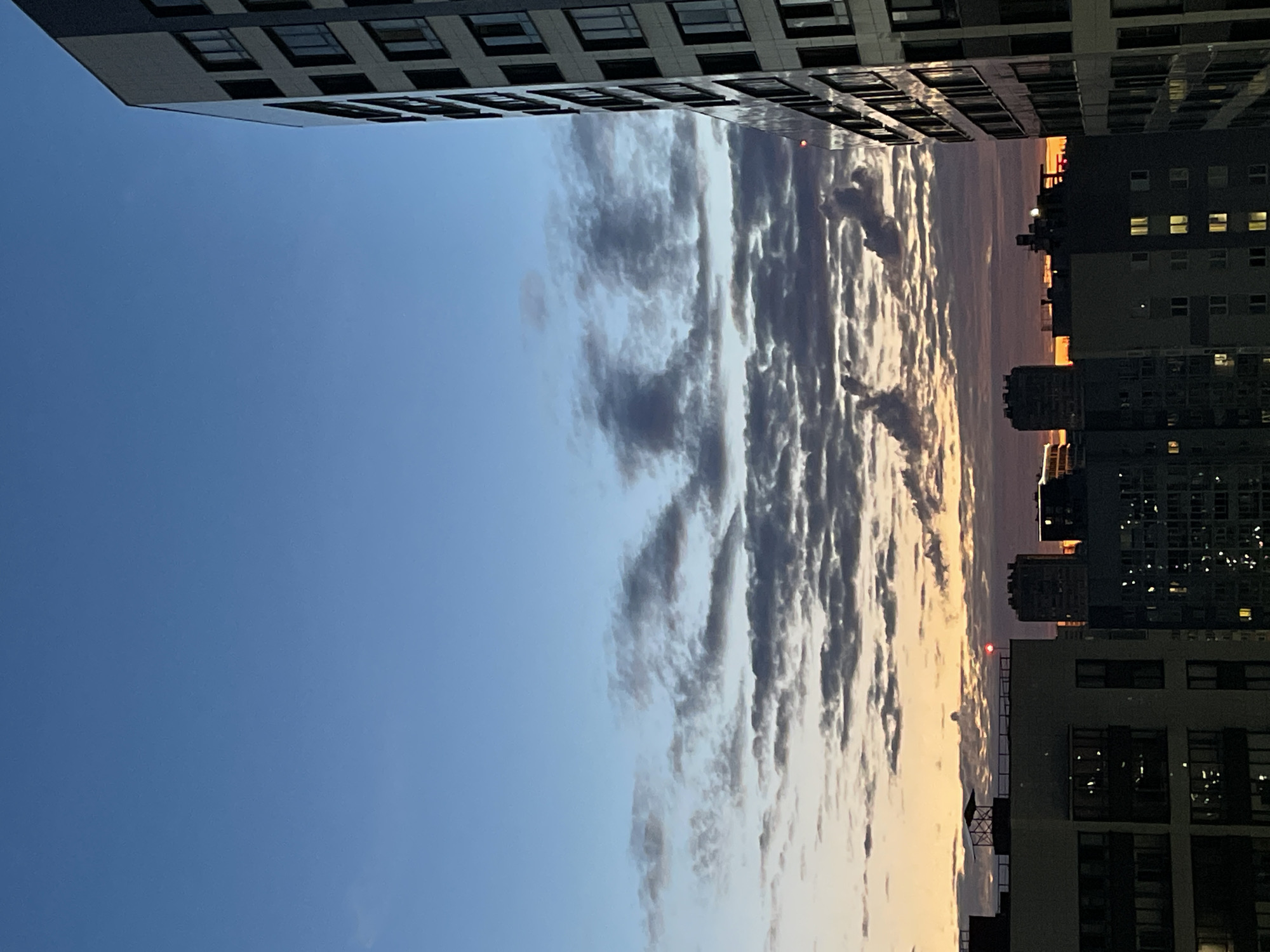 The media file is a low-angle, vertically oriented photograph of a cityscape at dusk. The scene is dominated by a clear blue sky on the left, transitioning to a gradient of orange and pink hues near the horizon on the right, indicating the setting sun. A band of scattered, dark clouds stretches across the mid-section of the image, illuminated from behind by the sunset.
Framing the sky on the left and right are the facades of tall apartment buildings. These buildings feature numerous windows, some of which are lit, suggesting occupancy. The architectural style appears modern, with repetitive geometric patterns of windows. On the far right, a smaller building with a red light on top is visible. The perspective of the photograph, looking upwards from ground level or a lower floor, emphasizes the scale of the buildings and the expanse of the sky.
The time of day is clearly dusk, given the colors of the sky and the illuminated windows in the buildings. The weather appears clear with scattered clouds. There are no visible people, but the lit windows imply human presence within the buildings. The location is identified as unknown, Russia, but the urban setting with apartment blocks is a common sight in many cities. The overall mood evoked is one of calm and transition, as day gives way to night in an urban environment. There is no visible text in the image.