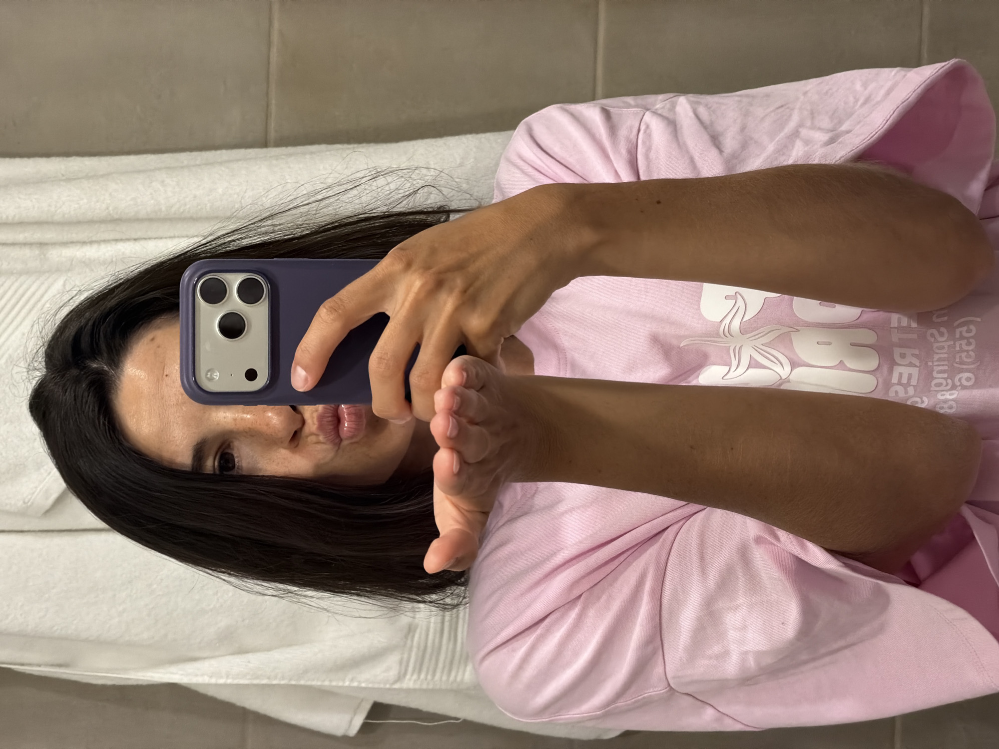 In a bathroom setting, a young woman with dark hair is taking a selfie with her phone. She is wearing a pink t-shirt with a graphic print on it. The woman is puckering her lips and blowing a kiss, holding her left hand up to the camera. A white towel hangs in the background, and the wall appears to be tiled. The image does not provide enough information to determine the specific location within l'Hospitalet de Llobregat, Spain.