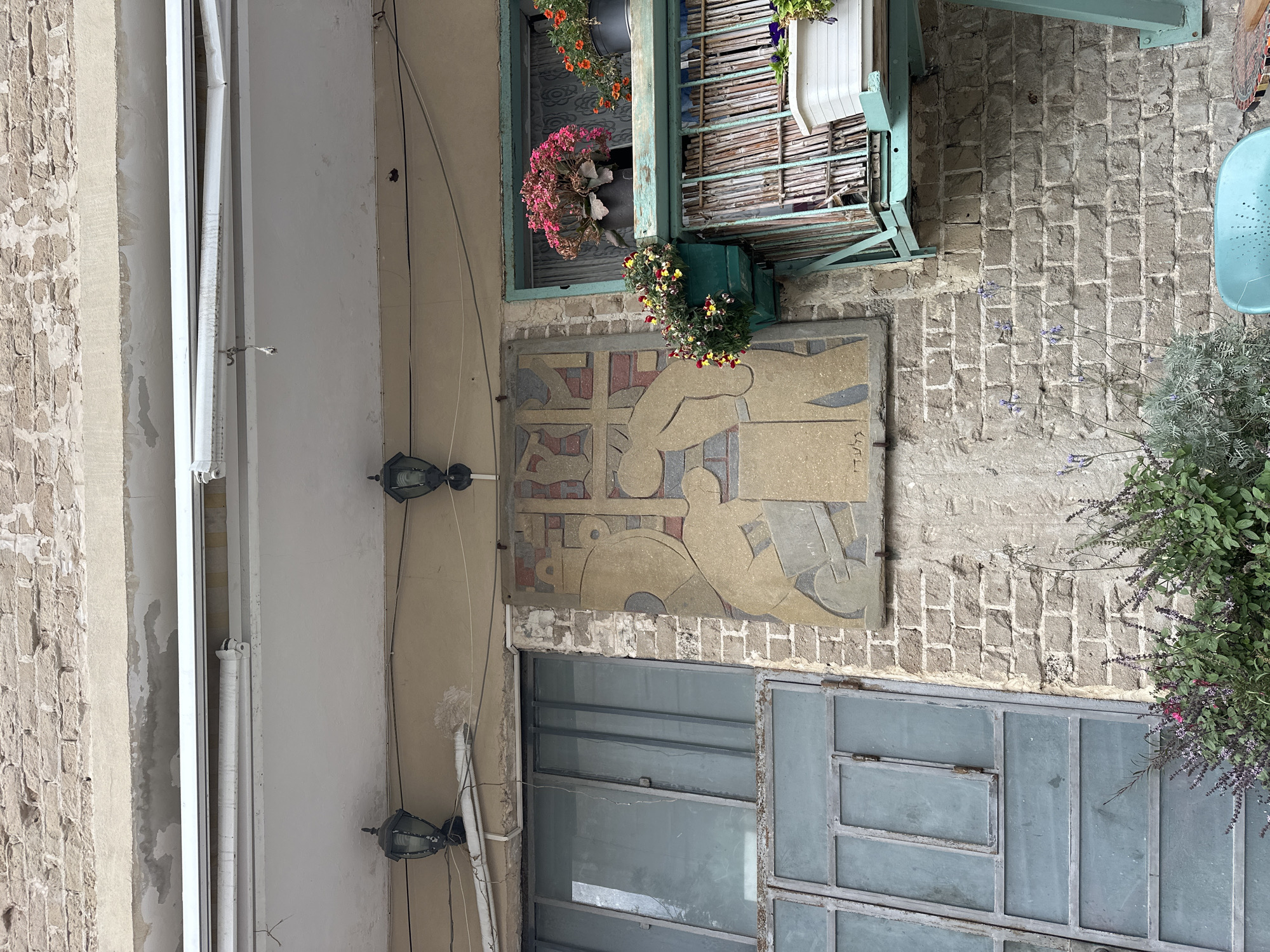 The image shows a vertical outdoor shot of a building in Tel-Aviv, Israel, with a brick and cement facade. A weathered artwork is affixed to the wall, depicting a hand reaching towards abstract shapes in muted tones. Above the artwork, a window box with colorful flowers adds a vibrant touch. Below it, a set of gray-framed glass panels serve as a window or door. Two black outdoor lights are mounted on the adjacent wall, with wires running along the surface. A plant with purple flowers is visible near a light blue outdoor chair in the right part of the frame. The architecture indicates a potentially historic or bohemian area of the city.