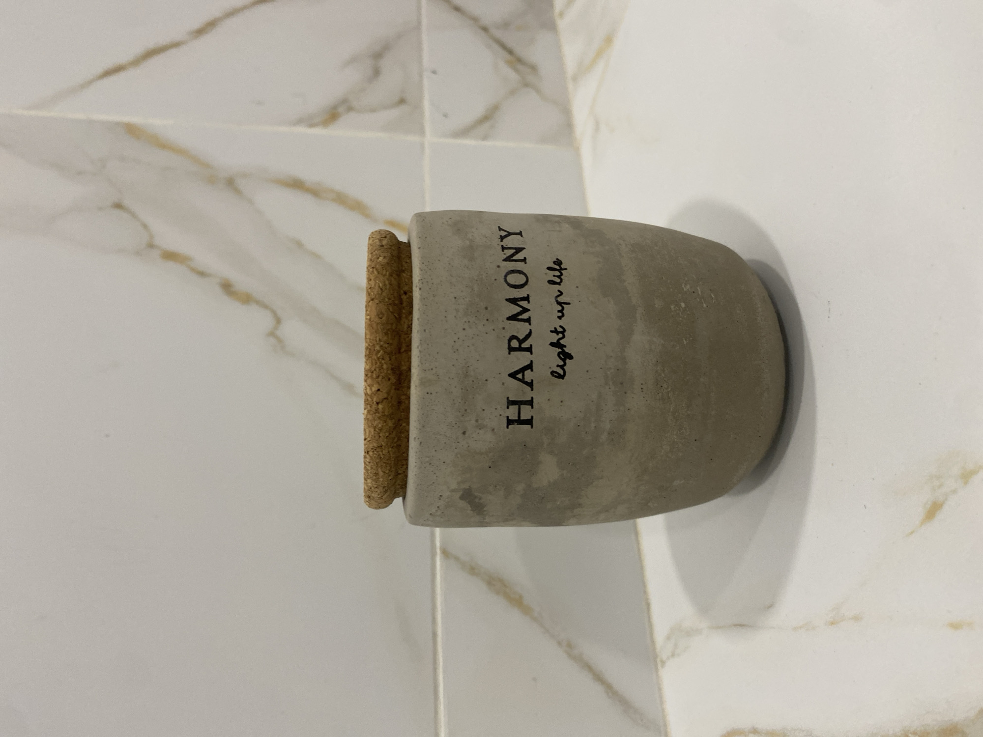 A close-up, eye-level shot shows a concrete candle holder with a cork lid placed on a white countertop with subtle gold veining. The candle holder is cylindrical with a slightly tapered base and features black text that reads "HARMONY" in a serif font, with "light up life" in a script font directly below. The background consists of a tiled wall with a similar gold-veined pattern. No people or animals are present in the scene. The lighting is even, and the focus is on the candle holder. This scene does not provide any specific information about its location in Chișinău, Moldova.