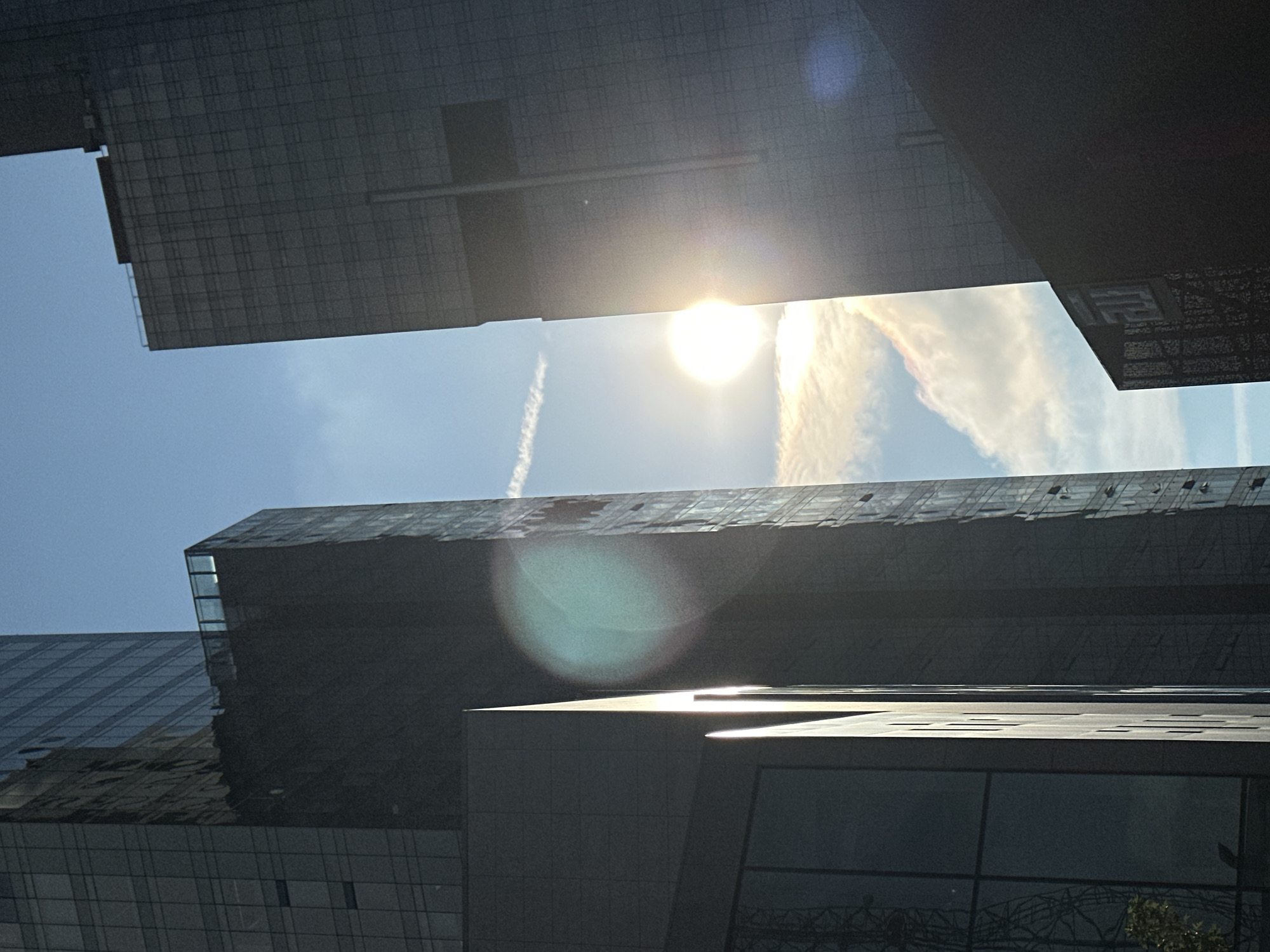 The image shows a low-angle shot looking up at modern glass-facade skyscrapers in the Şişli district of Istanbul, Turkey. The sun is shining brightly, creating lens flare and highlighting the edges of the buildings and some clouds in the sky. A contrail from an airplane is visible in the upper portion of the sky. The buildings are dark and angular, with many windows reflecting the light. One building in the foreground has a sharp, pointed edge where the light is most intense. The overall impression is one of urban grandeur and the starkness of architectural design against the natural elements. There are no people visible in the image.