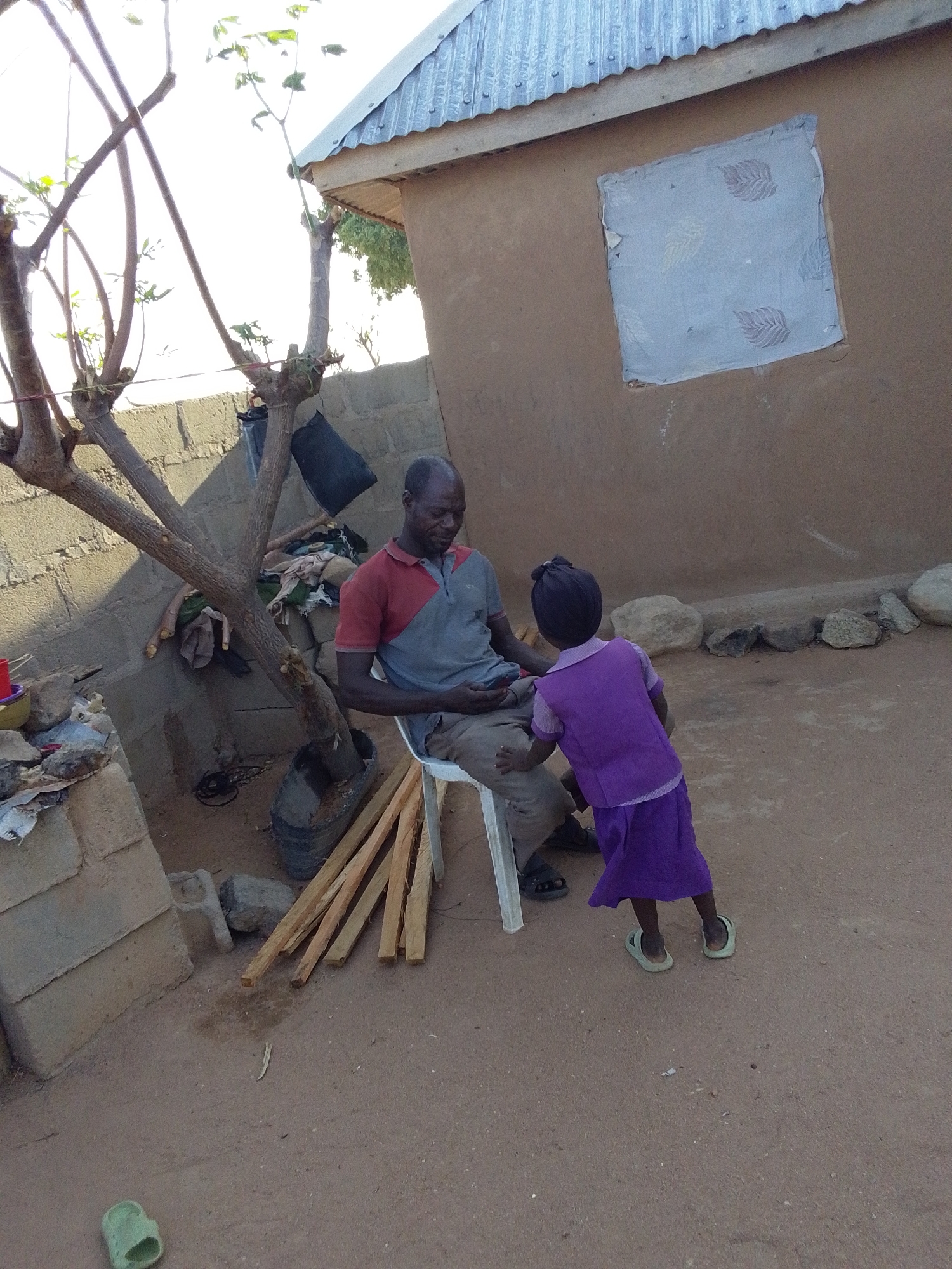 The image shows an adult male seated on a white plastic chair in an outdoor setting. He is wearing a two-tone shirt (red/grey) and light brown pants. A child in a purple dress is standing in front of the man, facing him. The surroundings feature a building with a corrugated metal roof and a window covered with a cloth featuring a leaf pattern. Adjacent to the building is a wall made of concrete blocks, and a bare tree is positioned near the seated man. There are wooden planks leaning against the chair and tree, as well as loose objects scattered on the ground. The ground appears to be dirt. The location is specified as Mulke Barikin Sayejo, Nigeria.