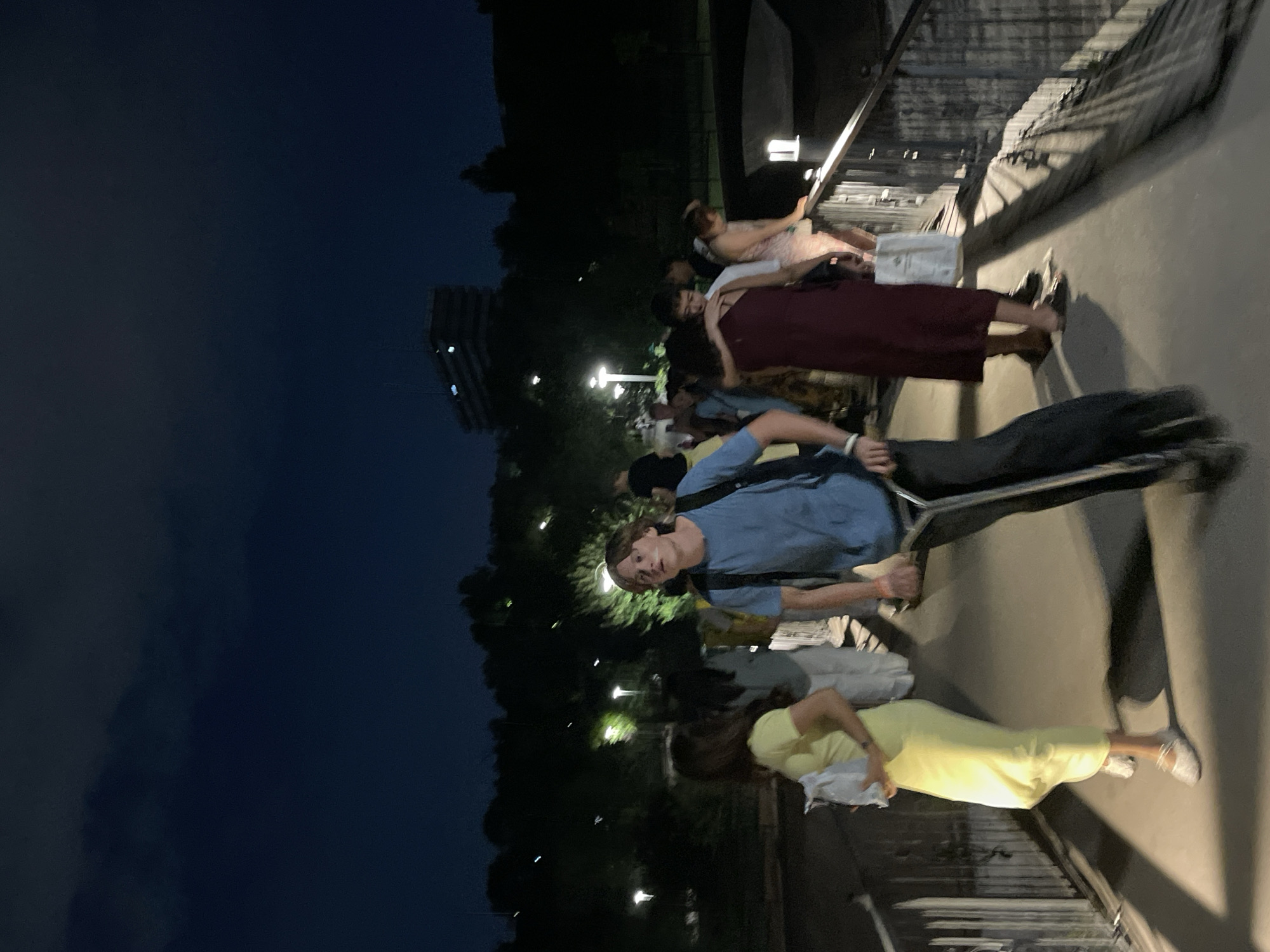 The image captures a nighttime scene on a pedestrian bridge, possibly in Almaty, Kazakhstan, given the context. The sky is dark blue, with some clouds. A young man is in the foreground, riding a scooter and looking upwards. He is wearing a blue t-shirt and dark pants, with a backpack on. To his left, a woman in a bright yellow dress is walking and holding a white bag. Several other people are visible on the bridge, walking in various directions. The bridge has metal railings and is illuminated by streetlights. In the background, there are trees and a building on a hill. The overall atmosphere is casual and public, suggesting an evening gathering or outing.