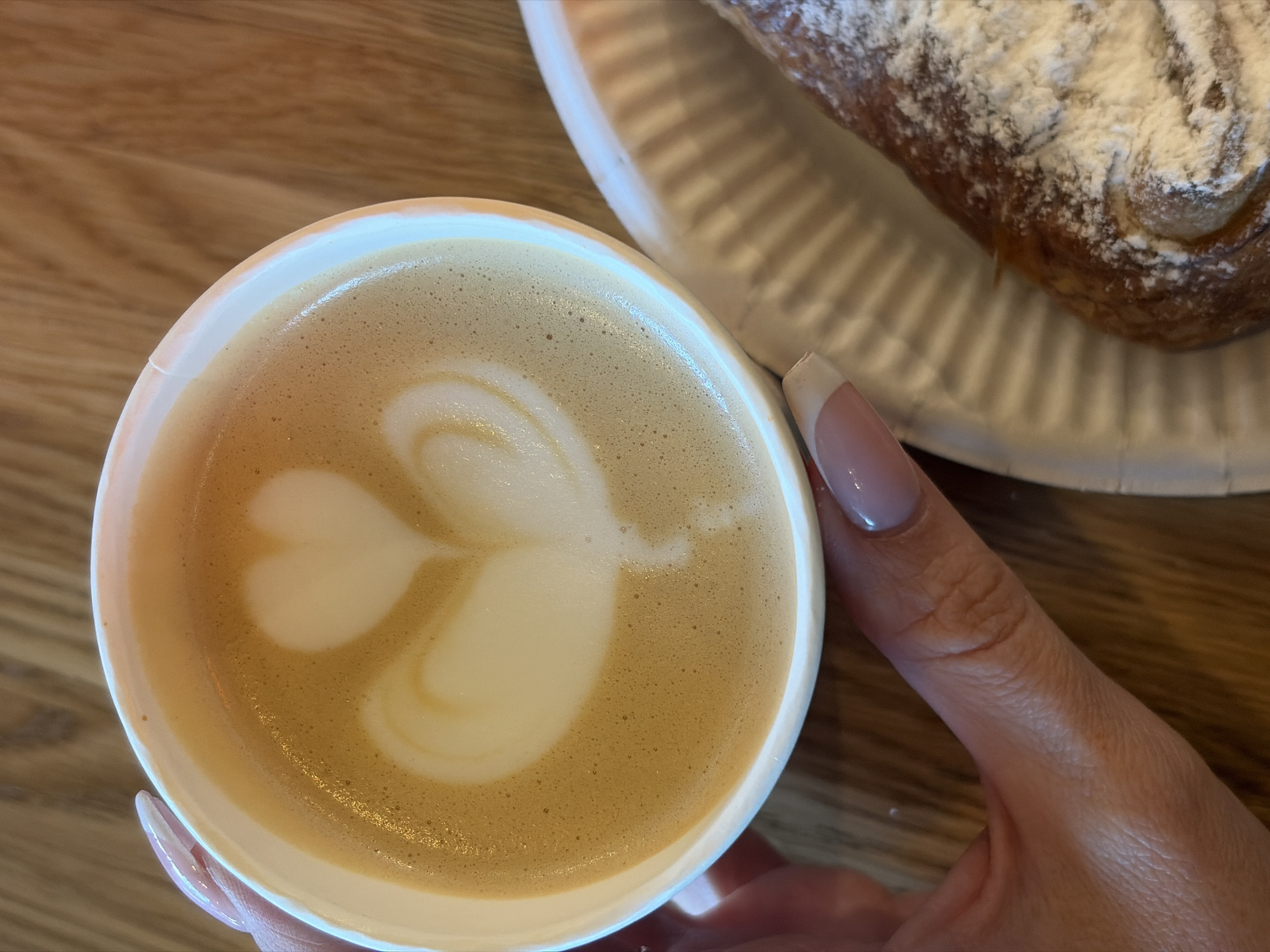 The image shows a close-up, top-down view of a cup of coffee held in a person's hand, alongside a pastry on a white plate. The coffee is a latte with latte art resembling a heart or tulip shape in the center of the foam. The cup is white, and the coffee appears to be light brown. The pastry is dusted with powdered sugar and is partially visible on the right side of the frame, sitting on a white paper plate with ridges. A fair-skinned hand with a long, manicured nail is holding the cup. The background is a light brown wooden surface. The location cannot be determined from the image.