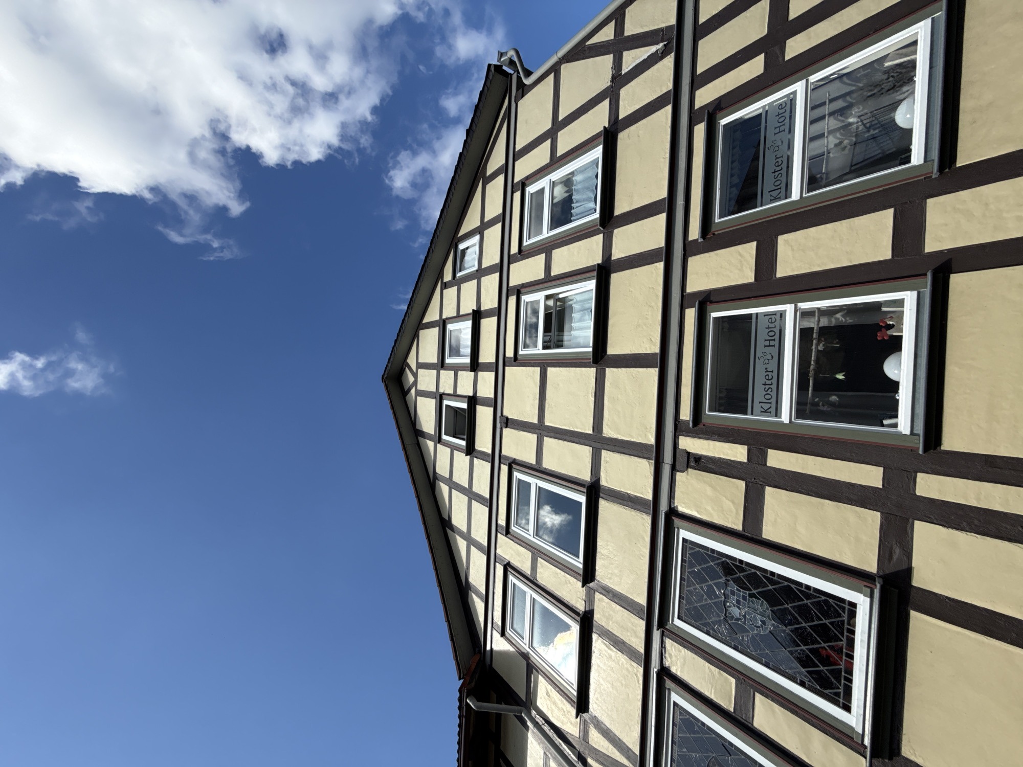 The image shows the facade of a historic half-timbered building with a steeply pitched roof against a bright blue sky with scattered white clouds. The building's exterior features a light yellow plaster with dark brown timber framing forming a grid pattern. Several windows of various sizes are visible on multiple levels. Some windows are plain with white frames, reflecting the sky and clouds. Others, particularly on the lower floors, have decorative stained glass or latticework patterns. One window on the upper floor has a sign that reads "Kloster Hotel." The perspective is from a low angle, looking upwards at the building. The setting appears to be a street in Walkenried, Germany, given the style of architecture often found in historic German towns. There are no people visible in the image.