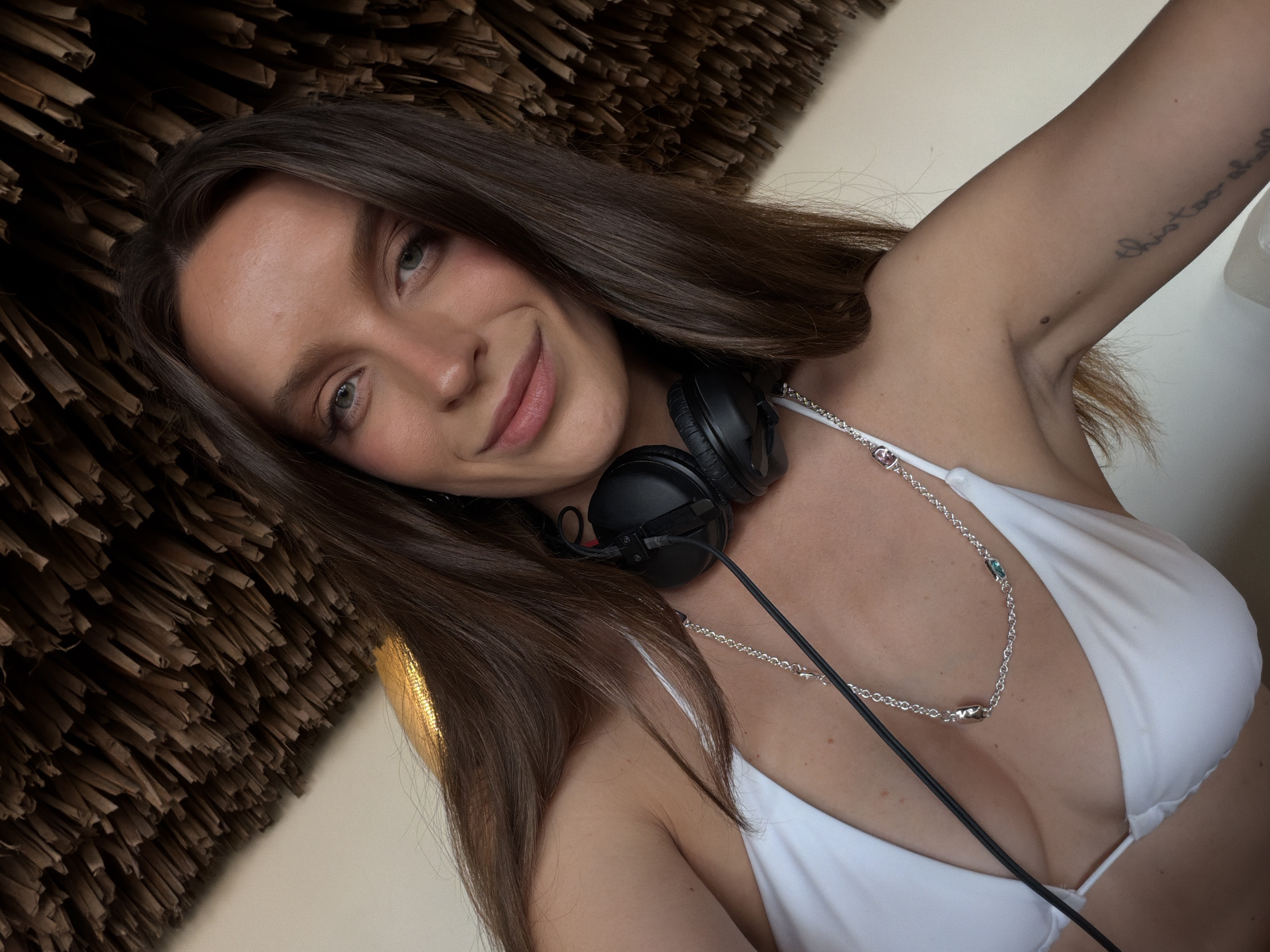 A woman with long, brown hair and a white bikini top is posing for a selfie. She is wearing black headphones around her neck and a silver chain with a pendant. Her left arm is tattooed with the phrase "this too shall pass." The background appears to be a thatched roof or wall. The image was taken in the city of Drobnići, Montenegro.