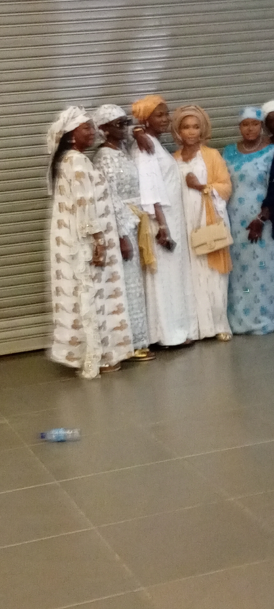 A group of five women, dressed in traditional Islamic attire, stand in front of a large, corrugated metal rolling door. The women are all wearing hijabs and long, flowing dresses. From left to right, the first woman wears a white dress with a gold patterned print and a white hijab. The second woman wears a grey and white patterned dress and a white hijab. She is wearing sunglasses. The third woman wears a white dress with an orange shawl draped over her shoulders, and a light brown hijab. She is carrying a tan shoulder bag. The fourth woman wears a white dress and a light blue hijab. The fifth woman, partially visible on the far right, appears to be wearing a blue and white patterned dress and a white head covering. In the foreground, on the tiled floor, lies a partially visible, clear plastic water bottle. The overall lighting suggests an indoor setting, possibly a public space or a community hall, with the rolling door indicating a closed or secured area. The image appears to have been taken in Gandu Albasa, Nigeria, a location known for its vibrant cultural attire and community gatherings.