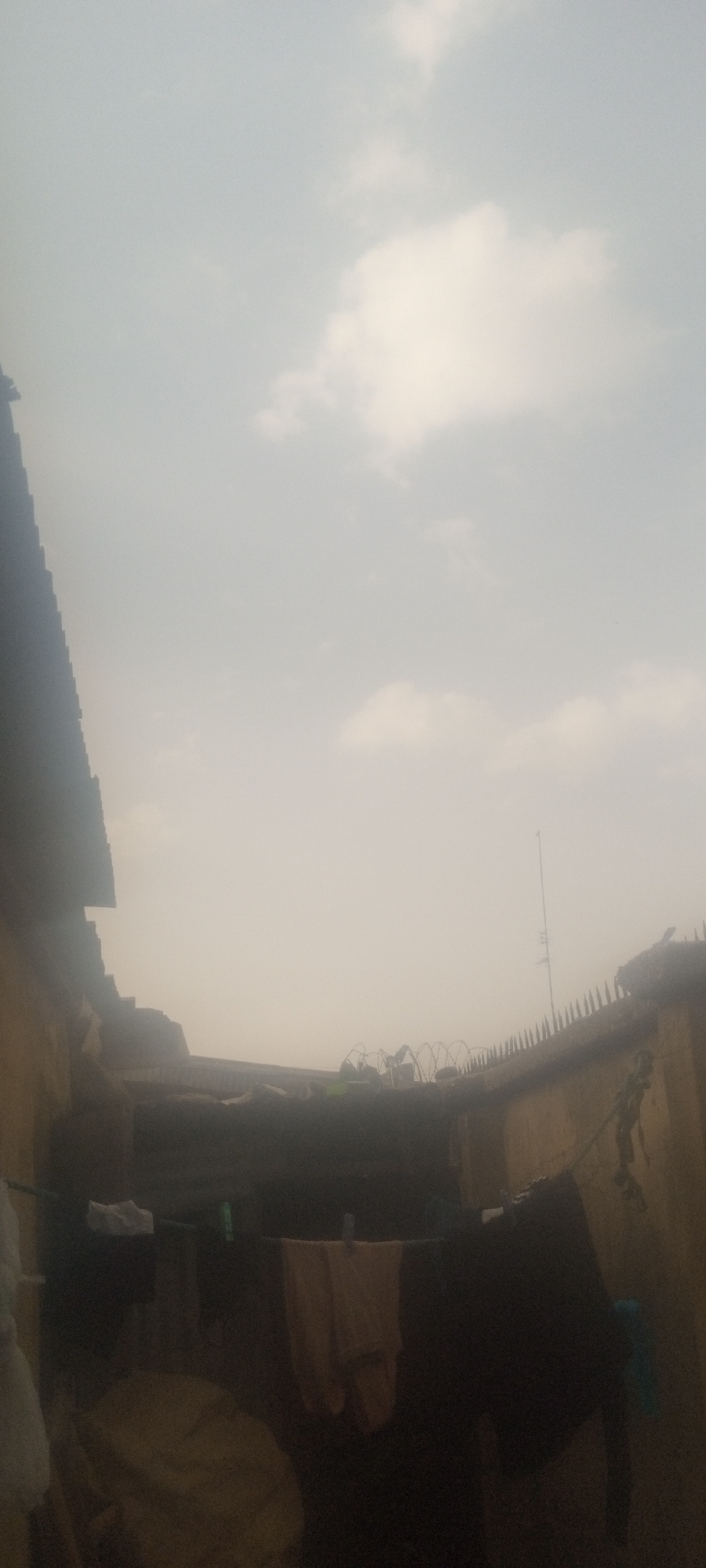 The scene depicts a rooftop view in Sabon Tasha, Nigeria. The sky is overcast with hazy white clouds. A corrugated metal roof is visible on the left. In the foreground, clothes hang on a line. The rooftop is secured with barbed wire and metal spikes. An antenna is on the right side of the image. The overall scene is dimly lit.