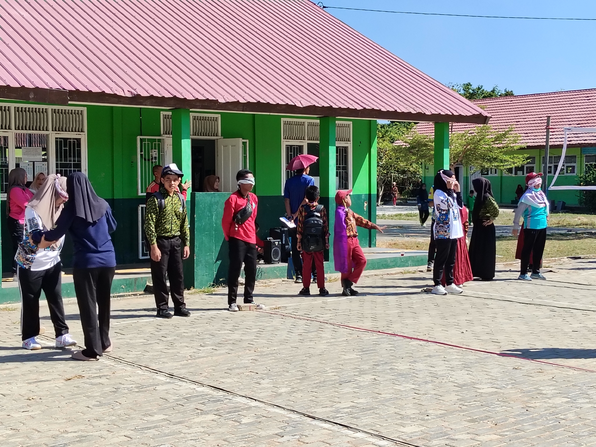 In Lori, Indonesia, on a sunlit day, an outdoor activity is underway in what appears to be a schoolyard with a green building featuring a pink roof. Several individuals are participating in an event that involves blindfolds. In the foreground, two young people, one in a dark blue top and black pants, and another in a white shirt with black pants, are standing close together. To their right, a person wearing a patterned green shirt and black pants is positioned slightly behind, with a blindfold covering their eyes. Further to the right, a person in a red shirt and black pants also has their eyes covered.
In the middle ground, a young person in a red shirt and black pants stands with a blindfold. To their right, a child in a patterned brown and orange shirt and red pants is facing away from the camera. A person in a purple shroud and a pink headscarf is also visible in this area. On the far right, a person in a light blue shirt and black pants is standing with their eyes covered. Behind them, other individuals are seen in the distance. A volleyball net is visible in the background, suggesting the presence of a sports court. The ground is paved with interlocking bricks.