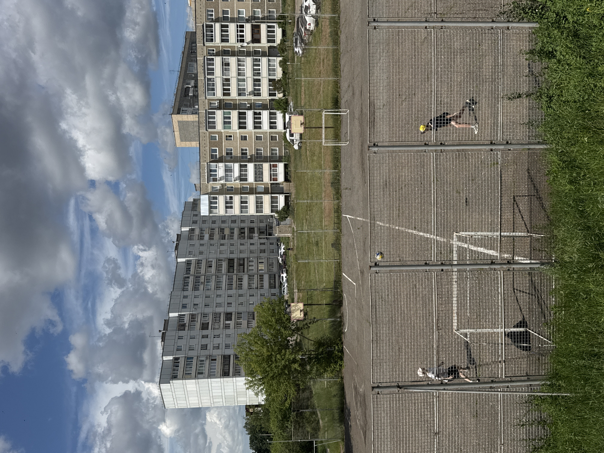 The image depicts an outdoor sports area with two young boys playing soccer. The area is enclosed by a chain-link fence. In the foreground, one boy wearing a white t-shirt and shorts is moving towards the left side of the frame. In the mid-ground, another boy in a black t-shirt and shorts with a yellow cap is seen moving towards the right. A soccer ball is in the air between them. A small soccer goal is visible in the foreground.
In the background, there are several apartment buildings, typical of Soviet-era architecture, and a basketball hoop. Cars are parked in front of the buildings, and some trees and grass are visible. The sky is partly cloudy, with dramatic clouds against a blue sky.
This scene is likely located in the city of Chaikovsky, Russia, given the architectural style of the buildings and the overall urban landscape.