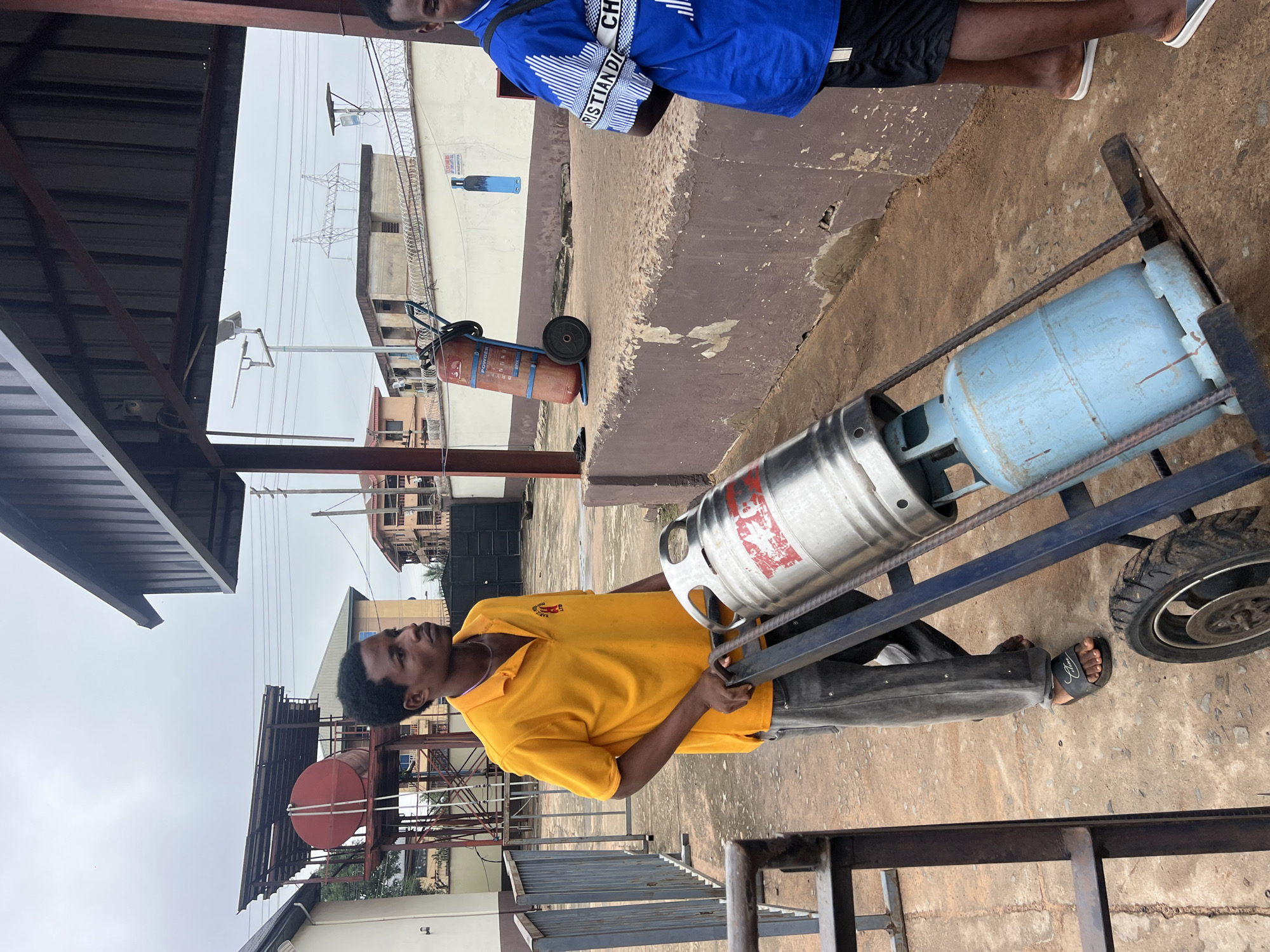 In Nkpor, Nigeria, a young man in a yellow polo shirt and grey trousers pushes a handcart loaded with a keg and a gas cylinder. The keg is silver, cylindrical, and has red markings. The gas cylinder is blue and strapped to the cart. Behind him, another man in a blue and white patterned shirt stands to the right. The scene appears to be an outdoor area with a concrete ground, a low brown wall, and a metal railing. Buildings and a large water tank structure are visible in the background. The sky is overcast.