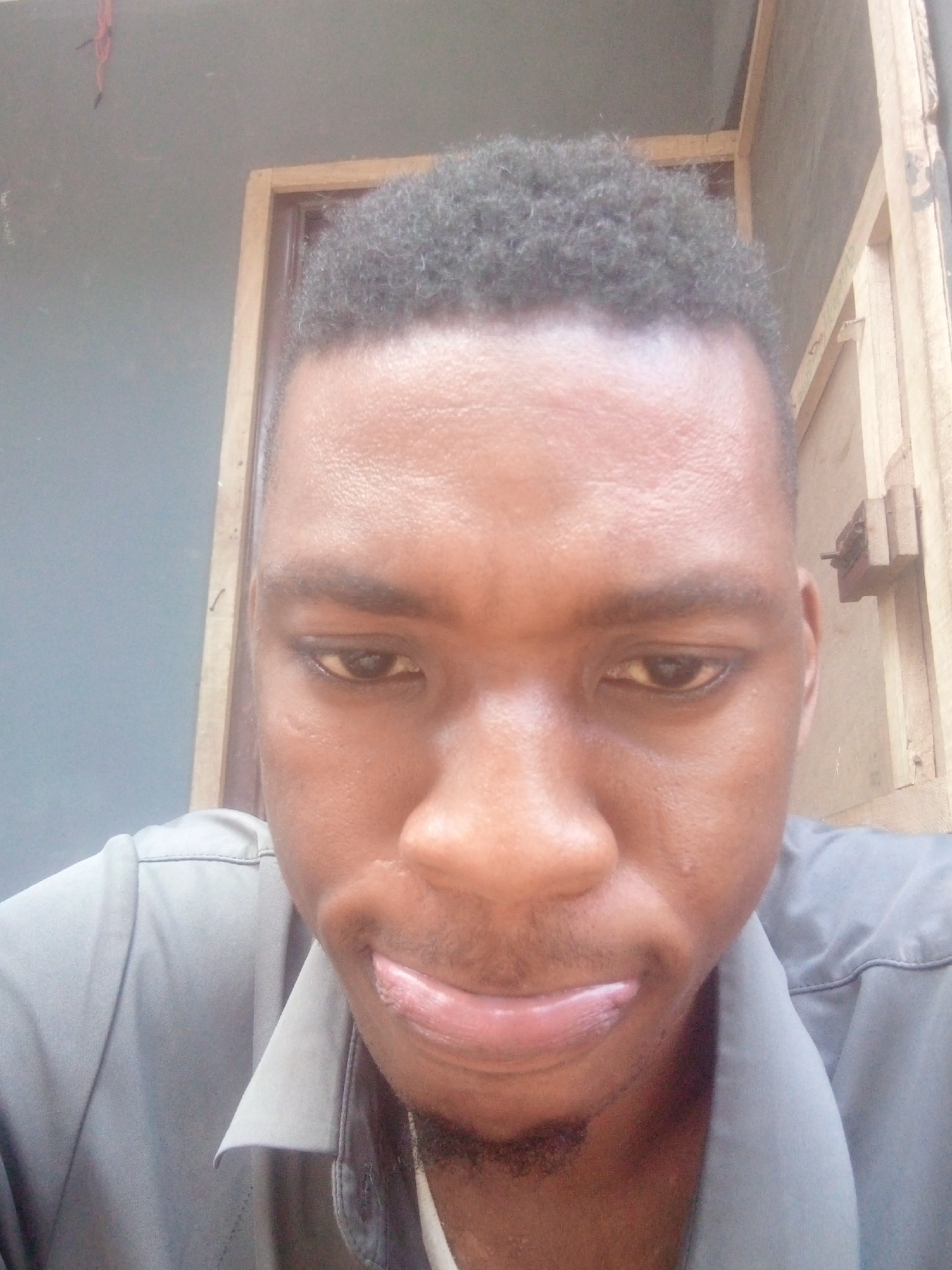 A medium close-up shot depicts a person with dark hair and brown skin, positioned in what appears to be an interior setting. The person's gaze is directed downward. Behind the individual is a wooden frame or door structure, and a gray wall is partially visible. The image suggests a domestic environment within the city of Ikorodu, Nigeria.