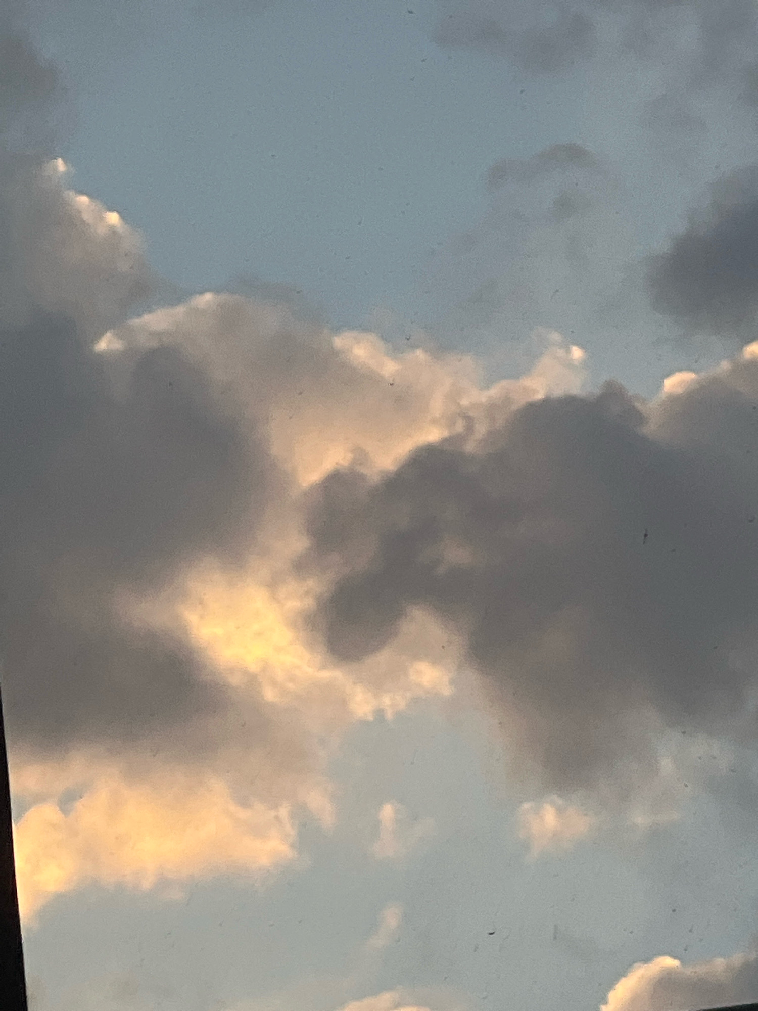A low-angle, medium shot captures a sky filled with clouds. The sky is a soft blue, with patches of light peeking through darker, grayer clouds. The clouds themselves are textured and voluminous, with some areas catching the sunlight and appearing golden or pinkish. The overall impression is one of a partly cloudy day, possibly at sunrise or sunset, given the warm tones in some of the clouds. There are no people, structures, or recognizable landmarks visible in the frame. The image appears to be taken from an outdoor perspective.