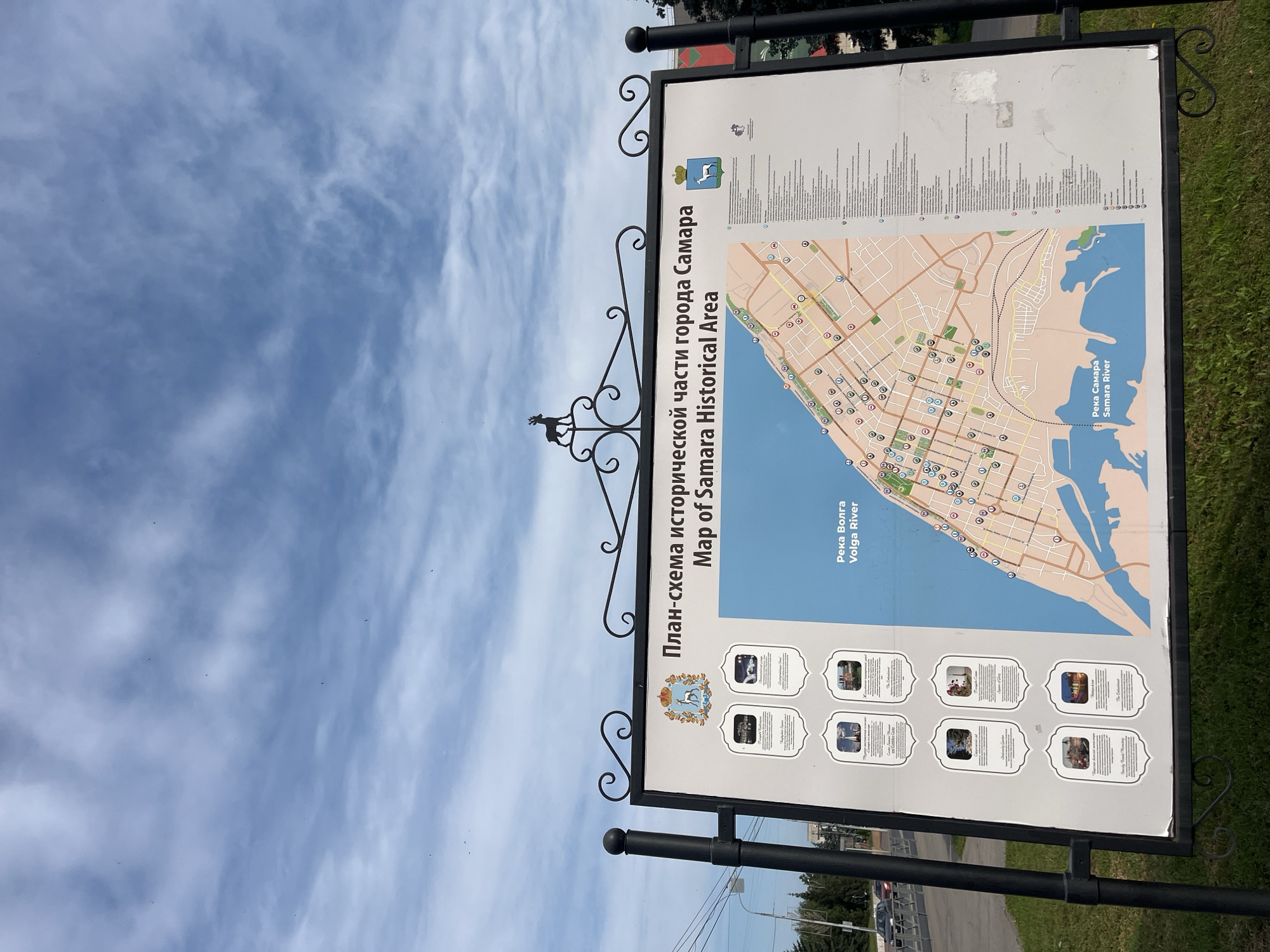 The image shows an outdoor information board in Samara, Russia, displaying a map of the city's historical area and the Volga River. The board is black metal framed with ornate scrollwork and topped with a silhouette of a deer. The map is on the right side of the board, and to the left are six framed images of notable landmarks with accompanying text in Russian. The sky above is cloudy, and the board is situated in a grassy area with a street and trees visible in the background.