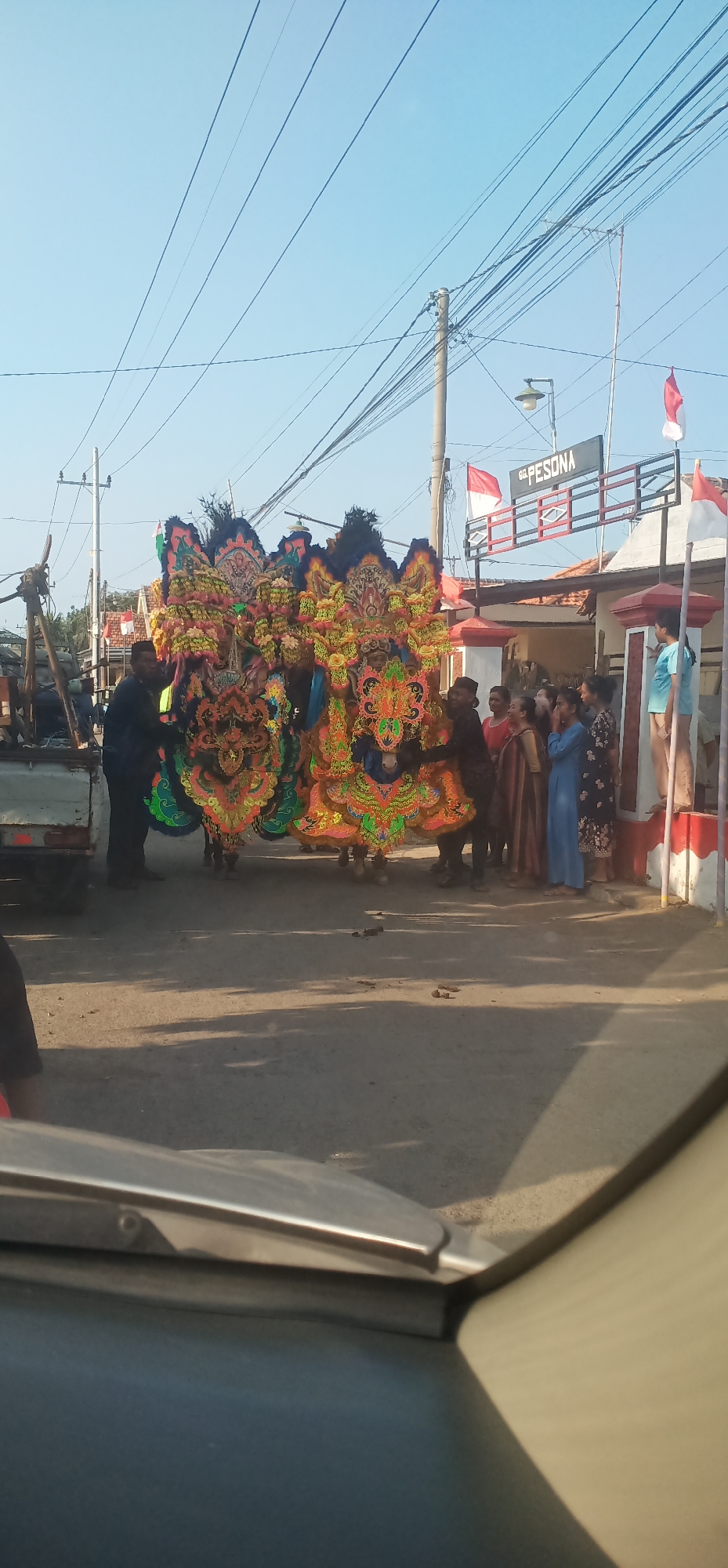 In Kalisalam, Indonesia, a procession features two large, ornate, dragon-like effigies, brightly colored in yellow, orange, green, and black. These figures appear to be mounted on a low platform or cart being pulled by unseen individuals or a vehicle, as two men in dark clothing are visible near the front of the effigies, seemingly guiding them. The scene is set outdoors on a paved road, with a clear blue sky overhead and visible power lines stretching across the frame. To the right, a building with a sign that reads "GG PESONA" is present, adorned with Indonesian flags (red and white). A group of spectators, including women in various colored clothing and at least one person in a light blue top, are gathered near the building, observing the procession. The foreground shows the hood and windshield of a vehicle, suggesting the image was taken from inside a car. The overall atmosphere appears to be festive, possibly part of a parade or cultural event.