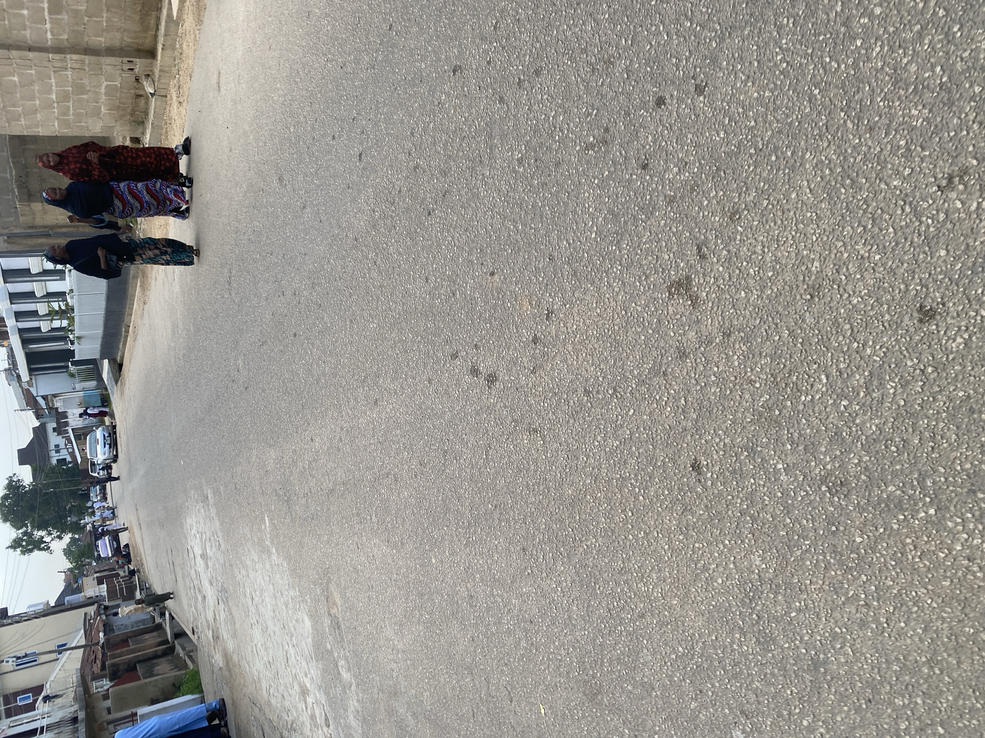 A street scene in Jos, Nigeria shows a paved road with buildings lining one side. Three women are walking together on the right side of the road. They are all wearing headscarves and long, patterned dresses. The woman on the far left wears a dark blue headscarf and a colorful, patterned dress. The middle woman has a black headscarf and a blue, red, and white patterned dress. The woman on the right has a red headscarf and a red and black patterned dress. They are walking away from the camera. Further down the street, a few people can be seen near parked cars. The sky is overcast.