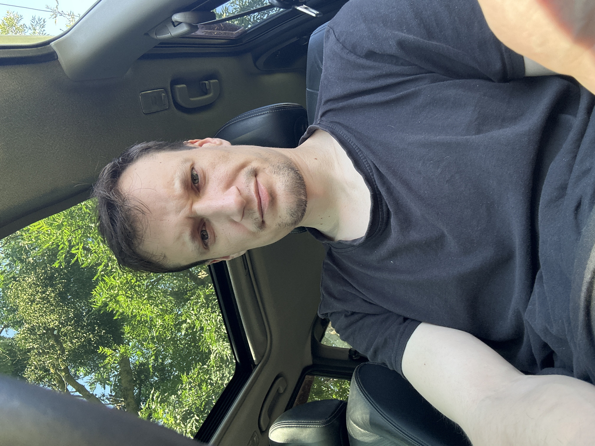 A man is sitting in the driver's seat of a vehicle, looking at the camera. He has short, dark brown hair and is wearing a black t-shirt. The sunroof of the vehicle is open, revealing green trees and a blue sky above. The interior of the car, including the dashboard and seats, is visible. The image appears to have been taken outdoors on a sunny day. The man is positioned in the center of the frame, and his body takes up most of the shot. The car is parked, as evidenced by the static nature of the scene.