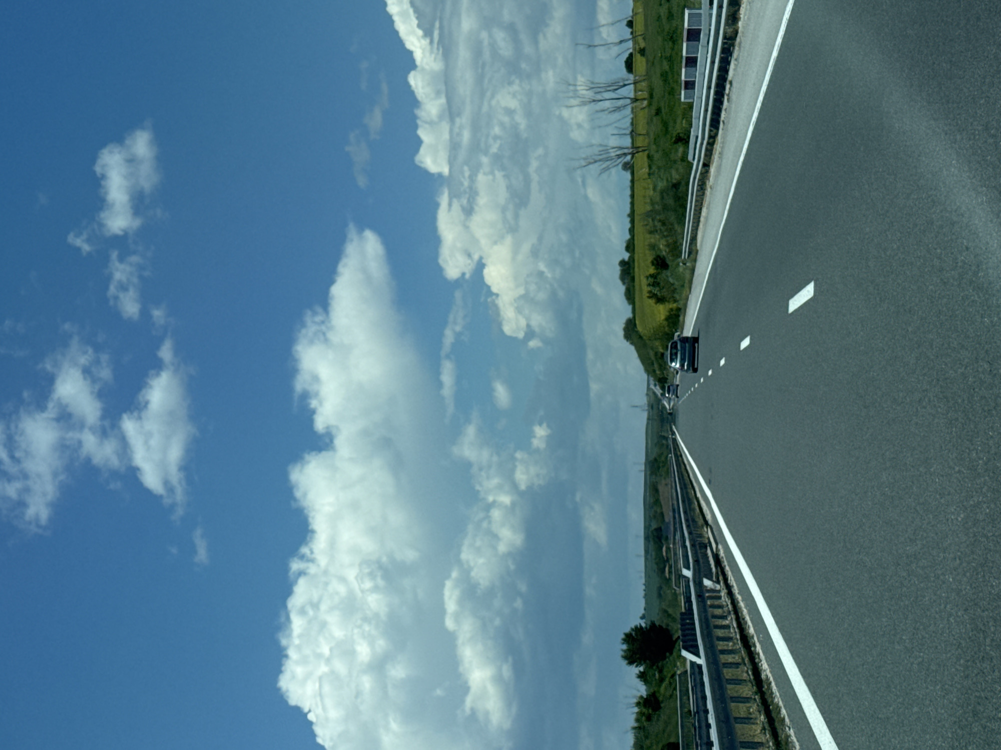 The image depicts a highway under a cloudy blue sky. The road is asphalt, with white lane markings and a guardrail on the left side. Vegetation lines the sides of the road. A dark-colored car is traveling in the same direction as the viewpoint. The location is in Panaiot-volovo, Bulgaria.