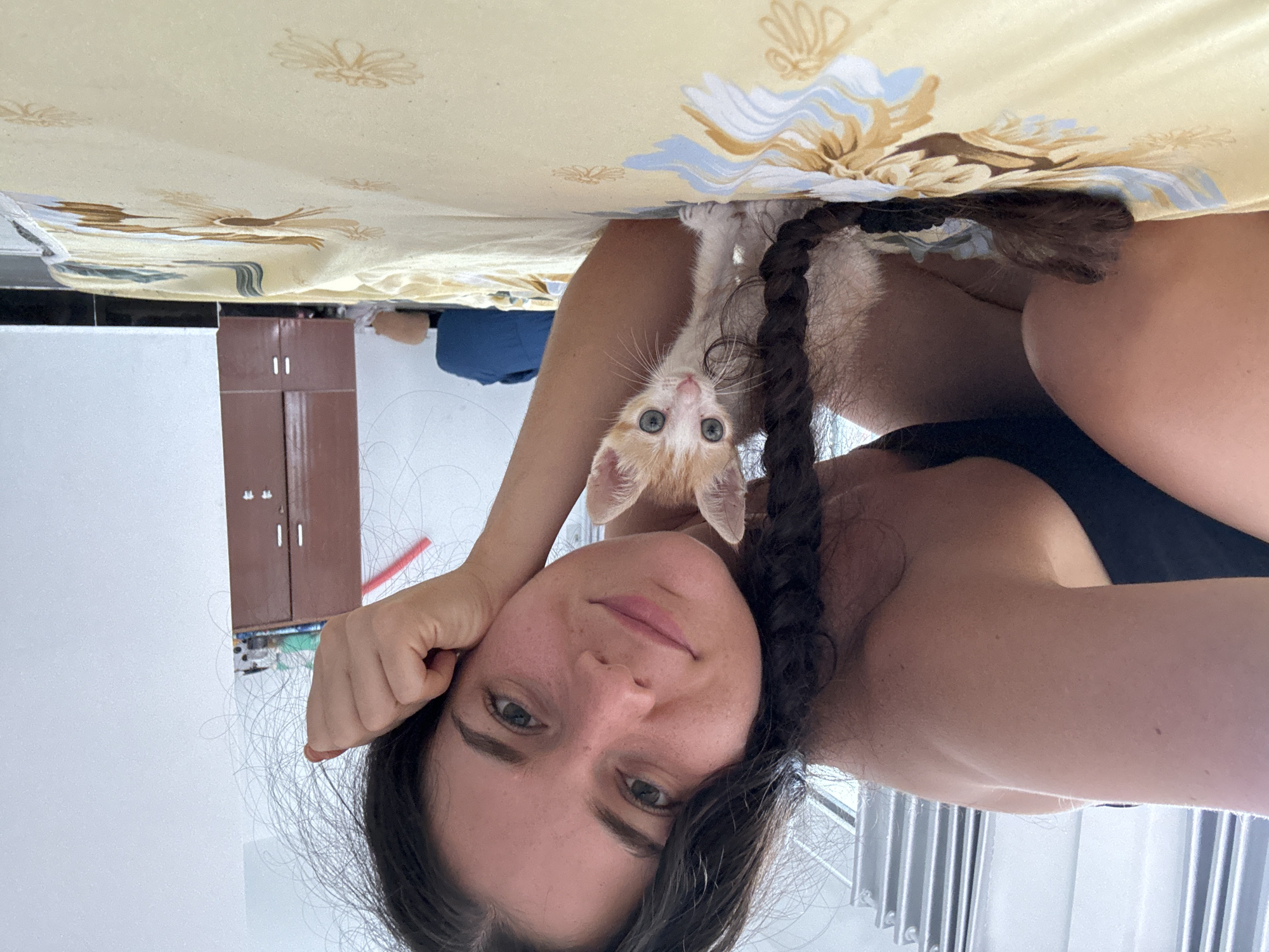 A person lies on their back with a small, orange and white kitten clinging to their chest. The person has dark hair, styled in a braid, and is looking directly at the camera with a slight smile. They are wearing a dark-colored top. The kitten is positioned between the person's face and chest. Above them is a patterned bedspread in shades of yellow and white. In the background, blurred, is a brown cabinet. The location is Mũi Né, Vietnam.