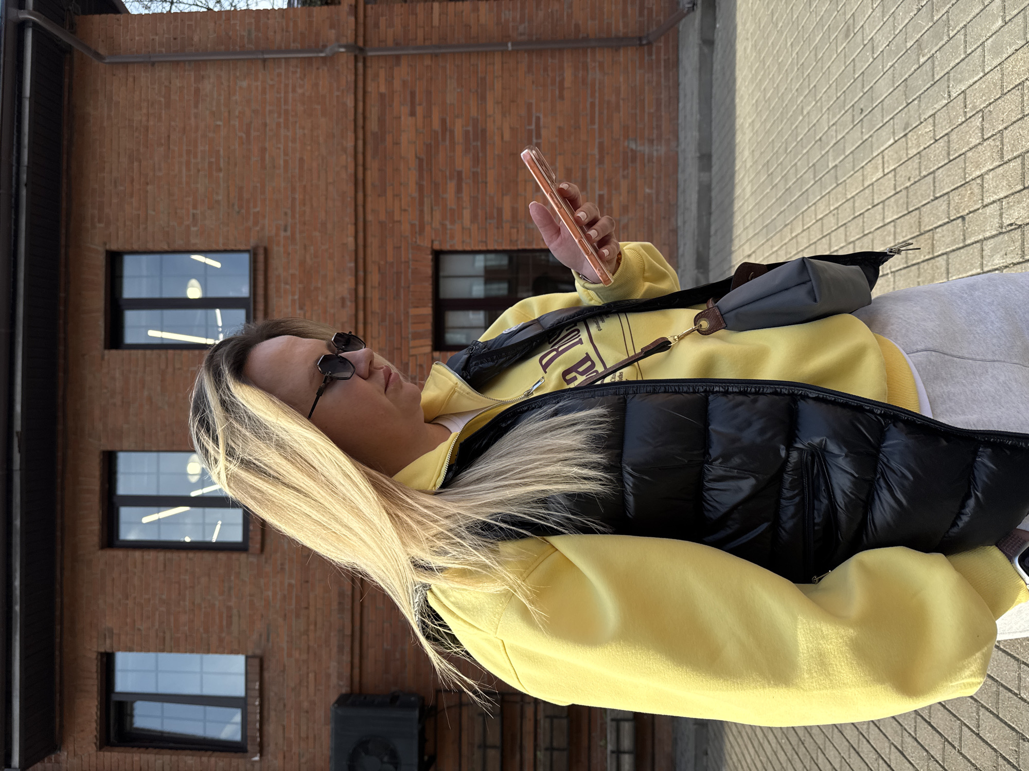 A woman with blonde-streaked hair, wearing sunglasses, a yellow sweatshirt, and a black vest, is standing outside a brick building while using a smartphone. She is also wearing a grey crossbody bag. The building features several windows and a black air conditioning unit. The ground is paved with light-colored bricks. This location is likely in the city of Moscow, Russia.