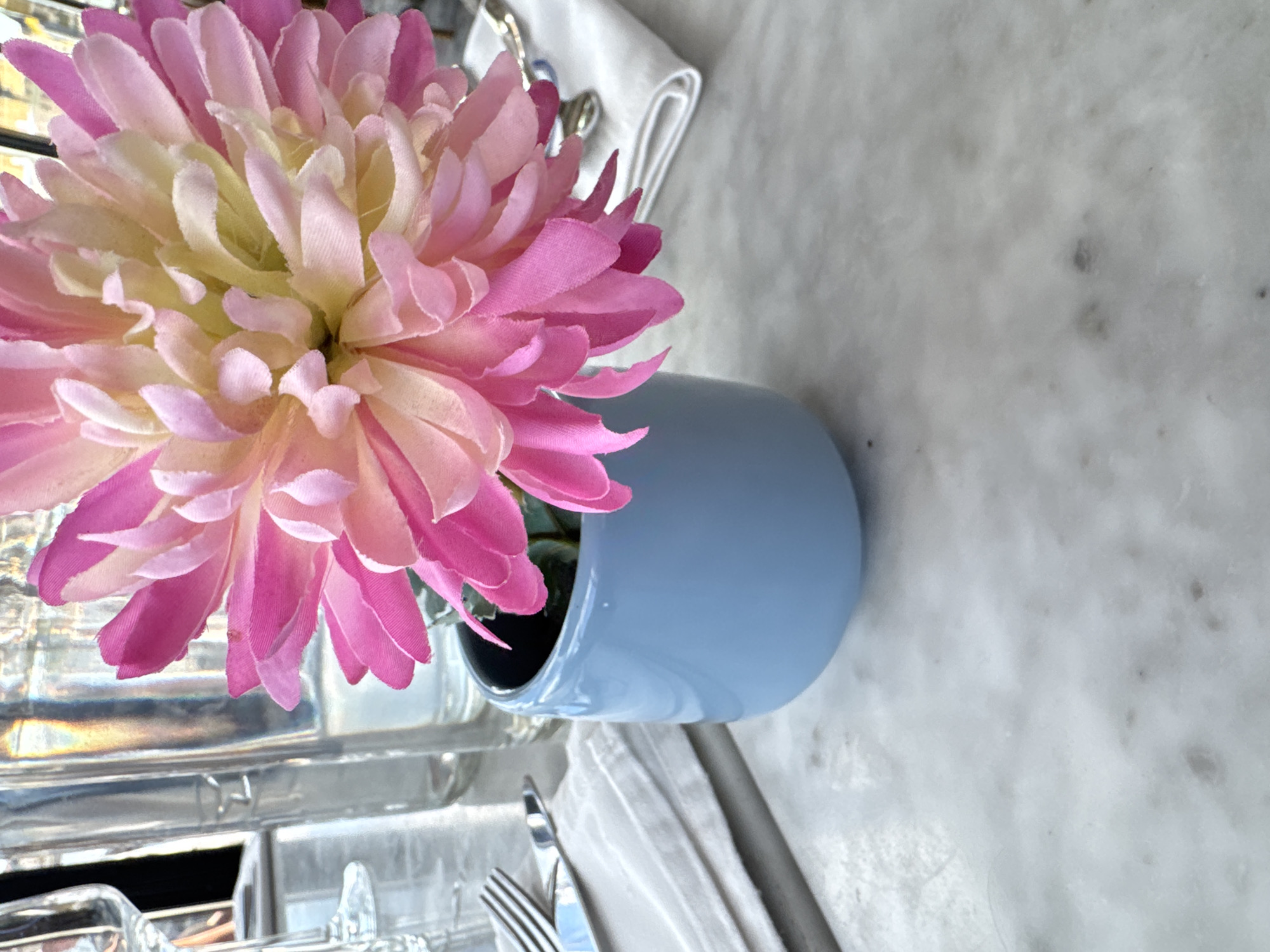 A close-up, eye-level shot shows a pink and yellow artificial flower with many petals in a light blue ceramic pot. The pot is situated on a white marble surface, with streaks of gray throughout. To the left of the pot, a folded white cloth napkin is visible, with a fork and knife resting on it. In the background, out of focus, are two clear glass bottles and a wine glass. The overall lighting is bright, with sunlight casting reflections on the glass bottles. The scene appears to be set on a table in an outdoor dining area.