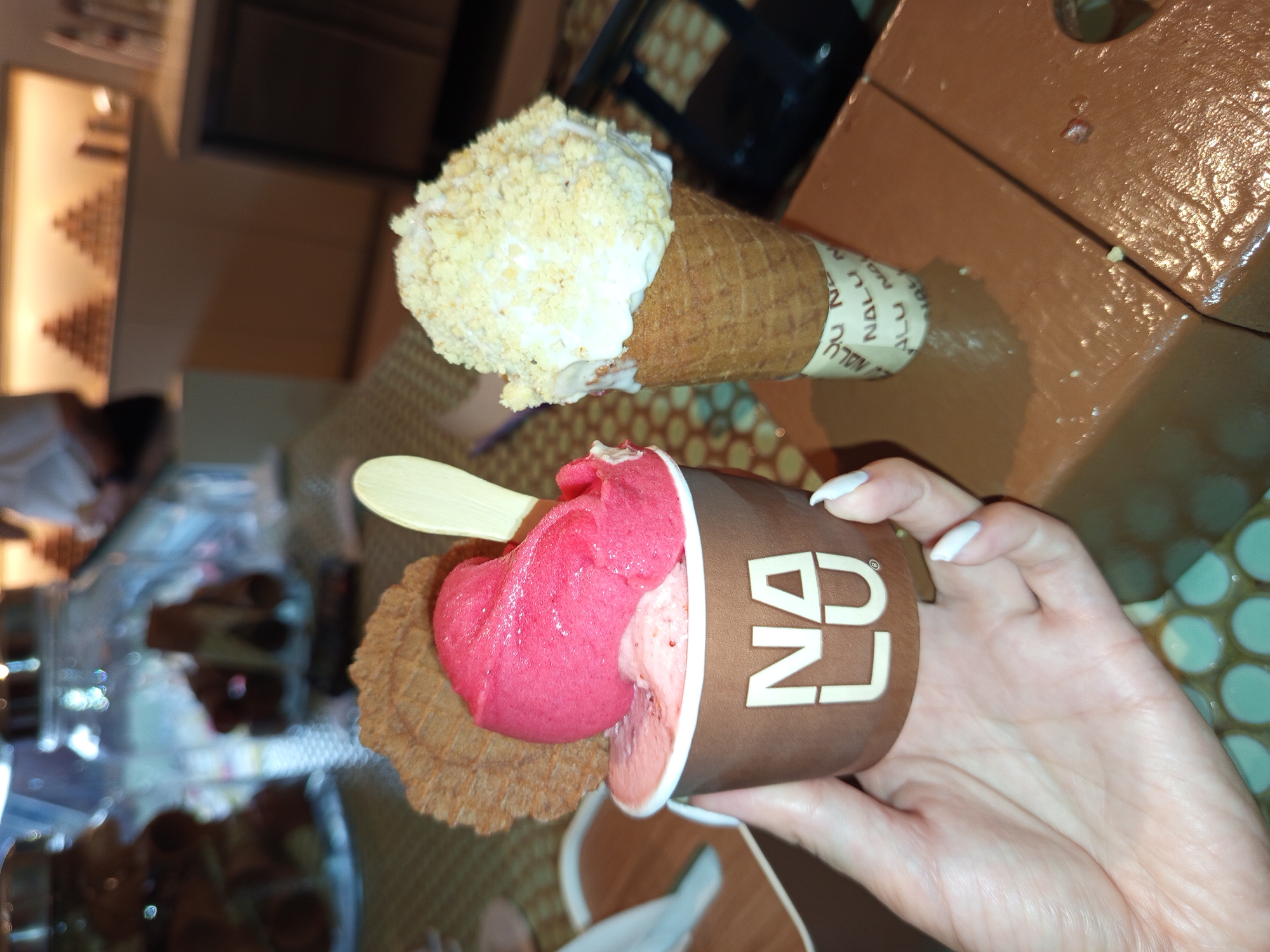 A person holds a cup of gelato and an ice cream cone. The cup contains pink and light pink gelato with a wafer sticking out. The ice cream cone contains white ice cream covered in crushed nuts. Both are from Nalu Bowls. The location is North Kuta, Indonesia. The background features a display case with pastries and a patterned table.