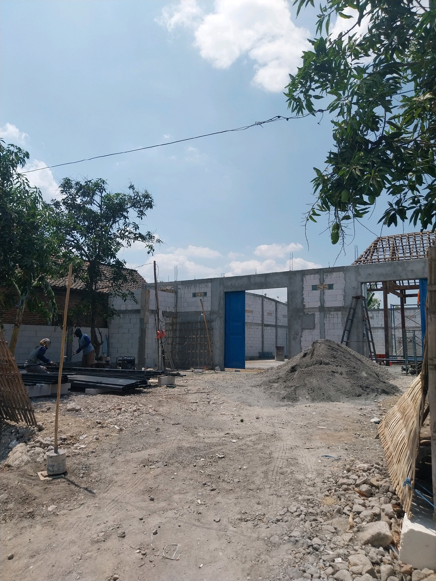 The image depicts an outdoor construction site in the city of Campur, Indonesia, under a clear blue sky with scattered white clouds. In the foreground, a dirt and gravel ground is visible with various debris and building materials scattered around. Two construction workers are present. One worker, wearing a blue shirt and light-colored pants, is standing and appears to be working on something. Another worker, in a grey shirt and dark pants, is crouching and also engaged in work. In front of them, a stack of long, dark metal pipes is neatly arranged on the ground, supported by concrete blocks. A tall, thin wooden pole stands vertically in the foreground, anchored by a concrete base.
In the midground, the construction of a building is underway. The structure is partially complete, with walls made of concrete blocks. A large blue door is visible, set into one of the walls. A pile of grey sand or cement is amassed near the building. A ladder leans against a partially constructed section of the building. Further in the background, a roof structure with wooden beams is visible, indicating ongoing construction. Trees with green leaves are present on the left and right sides of the image, with one tree in the upper right corner having visible green fruit hanging from its branches. The overall scene suggests an active construction project in a semi-urban or rural setting within Campur, Indonesia.