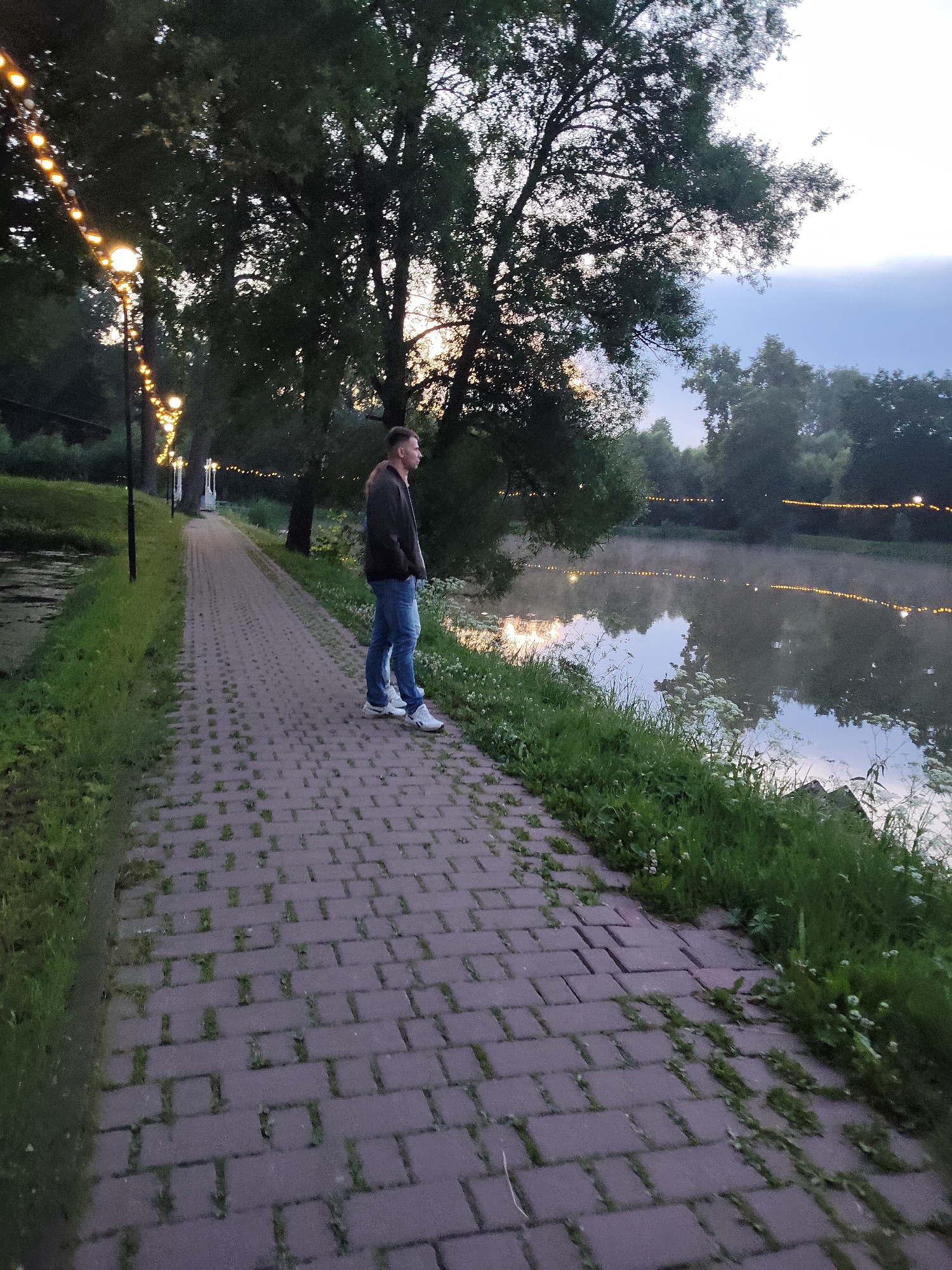 A man stands on a paved walkway next to a lake at dusk. The walkway is made of red brick pavers and is bordered by grass and foliage. String lights are visible in the background, illuminating the trees and the lake, which has a light mist rising from its surface. The man is wearing a dark jacket, blue jeans, and white sneakers. He is looking out at the lake. The scene appears to be a park or recreational area.