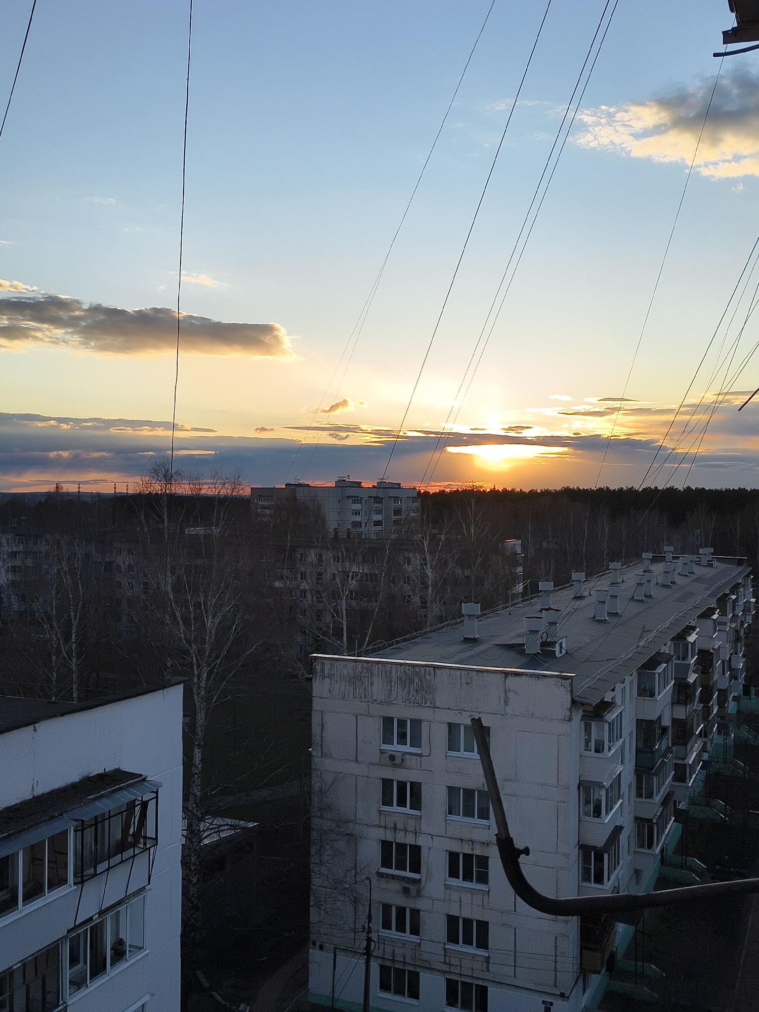 The image captures an urban landscape at sunset, viewed from a high vantage point, possibly from another building. The sky is a prominent feature, transitioning from a clear light blue at the top to a warm orange and yellow near the horizon where the sun is setting. Scattered clouds, some illuminated by the golden light, are visible.
In the immediate foreground, several thin, dark lines, likely wires or cables, crisscross diagonally across the upper portion of the frame. On the lower left, part of a white residential building with multiple windows and a balcony is visible. On the lower right, a large, dark, curved pipe or conduit extends upwards from the bottom edge.
The midground features numerous multi-story residential buildings, appearing to be concrete panel apartment blocks, with flat roofs. One large building in the center-right has several white ventilation units on its roof. Many bare trees, suggesting late autumn or early spring, are interspersed among the buildings, particularly towards the left and center. The buildings and trees are largely in shadow, creating a stark contrast with the brightly lit sky.
In the background, a dense line of trees, forming a forest, stretches across the horizon. Beyond this tree line, the setting sun casts a strong, radiant glow, with rays of light occasionally breaking through clouds. Distant, indistinct structures can be vaguely seen on the horizon line above the trees. No people or explicit activities are visible, and there is no discernible text in the image. The overall impression is one of a quiet, somewhat industrial or residential scene at dusk.
