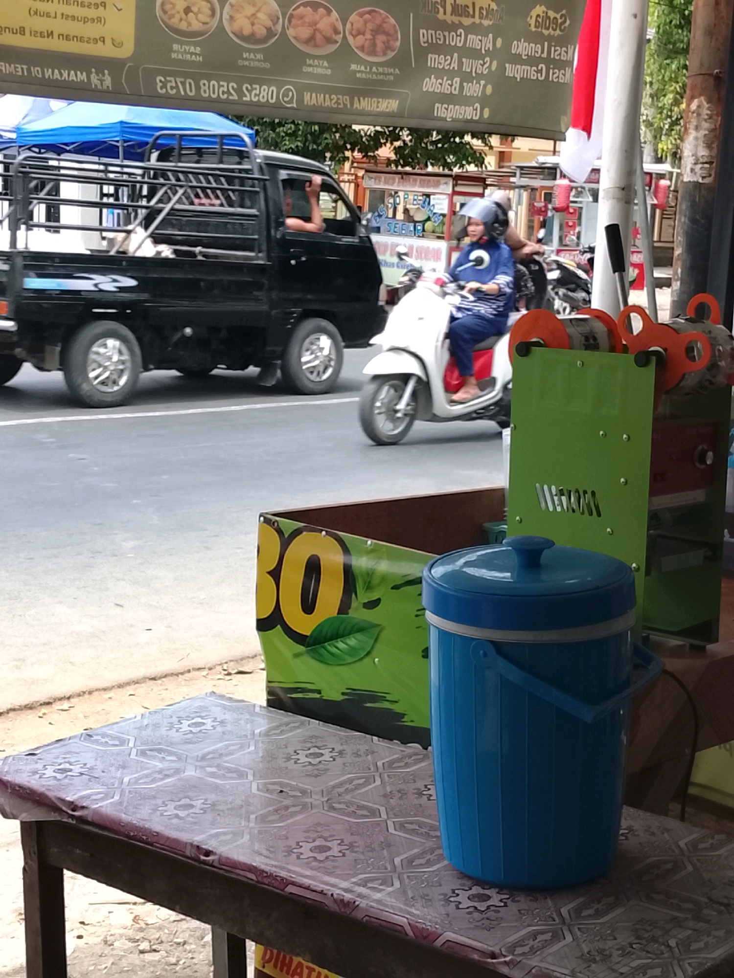 In Slawi, Indonesia, a black pickup truck drives on a paved road. A person with a raised arm is visible in the driver's side of the truck. In the background, a person on a white scooter wears a blue outfit and a helmet. A green machine with orange accents stands beside the road. A blue insulated jug sits on a table covered with a patterned cloth. A banner displaying the number "30" and a green leaf is partially visible behind the jug.